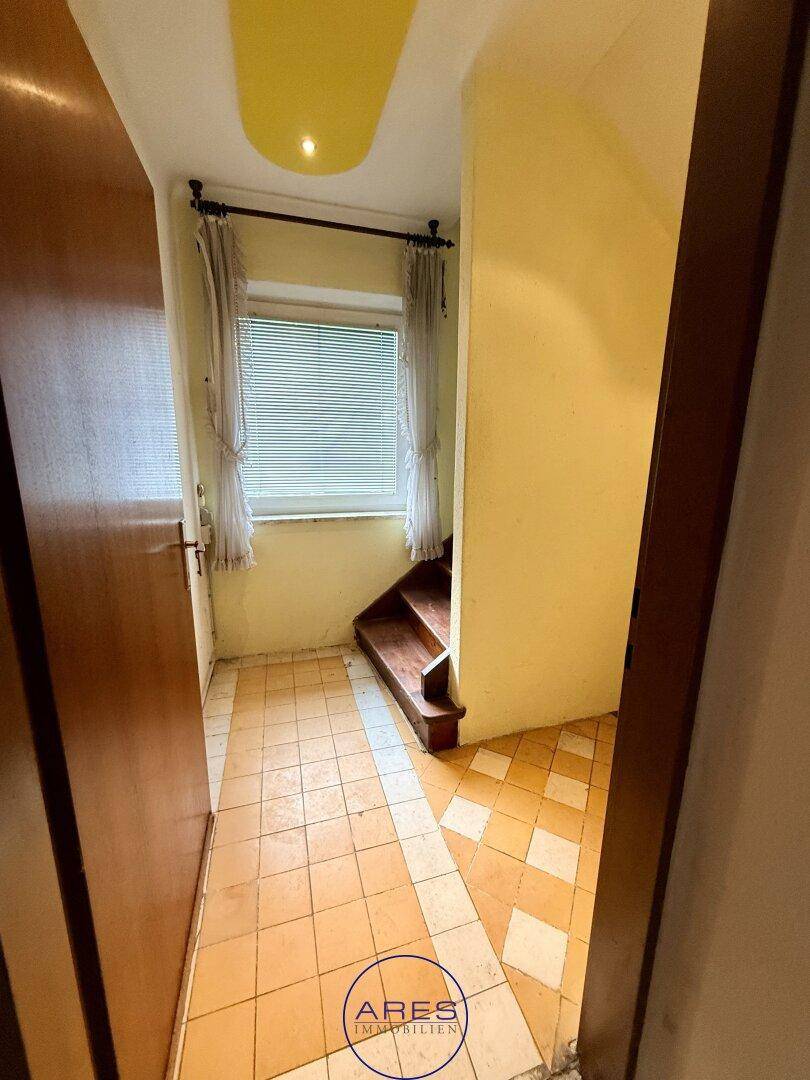 Flur mit Fliesenboden und Fenster, von dem eine Holztreppe in den Kellerbereich führt.