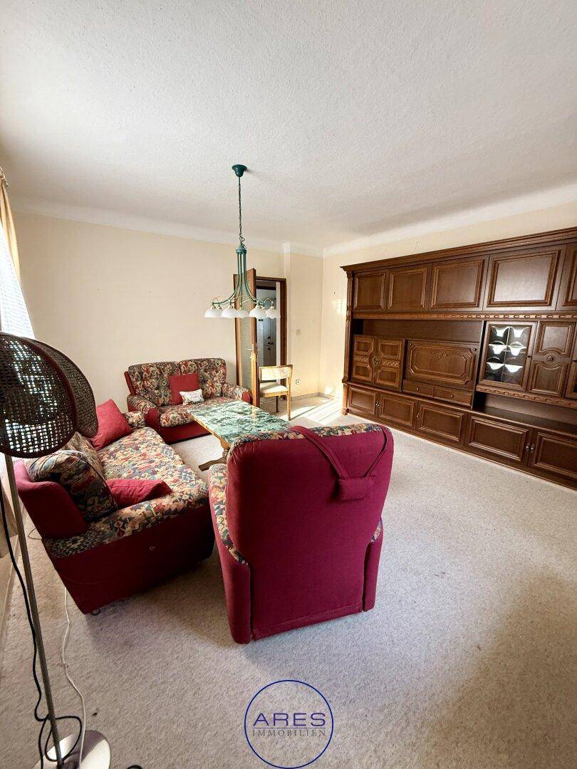 Geräumiges Wohnzimmer mit Teppichboden, großer Holzschrankwand und traditionellen roten Polstermöbeln.