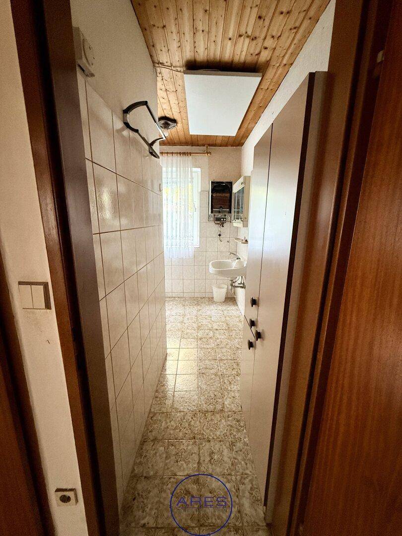 Badezimmer mit gefliestem Boden und Wänden, Waschbecken und Fenster, ergänzt durch eine Holzdecke.
