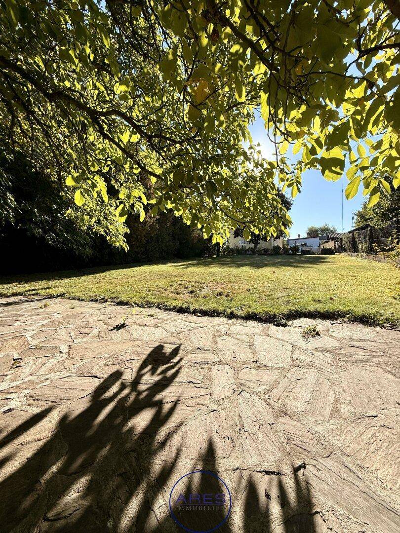 Steinplattenweg im sonnigen Garten mit weitem Rasen und schattenspendenden Bäumen im Hintergrund.