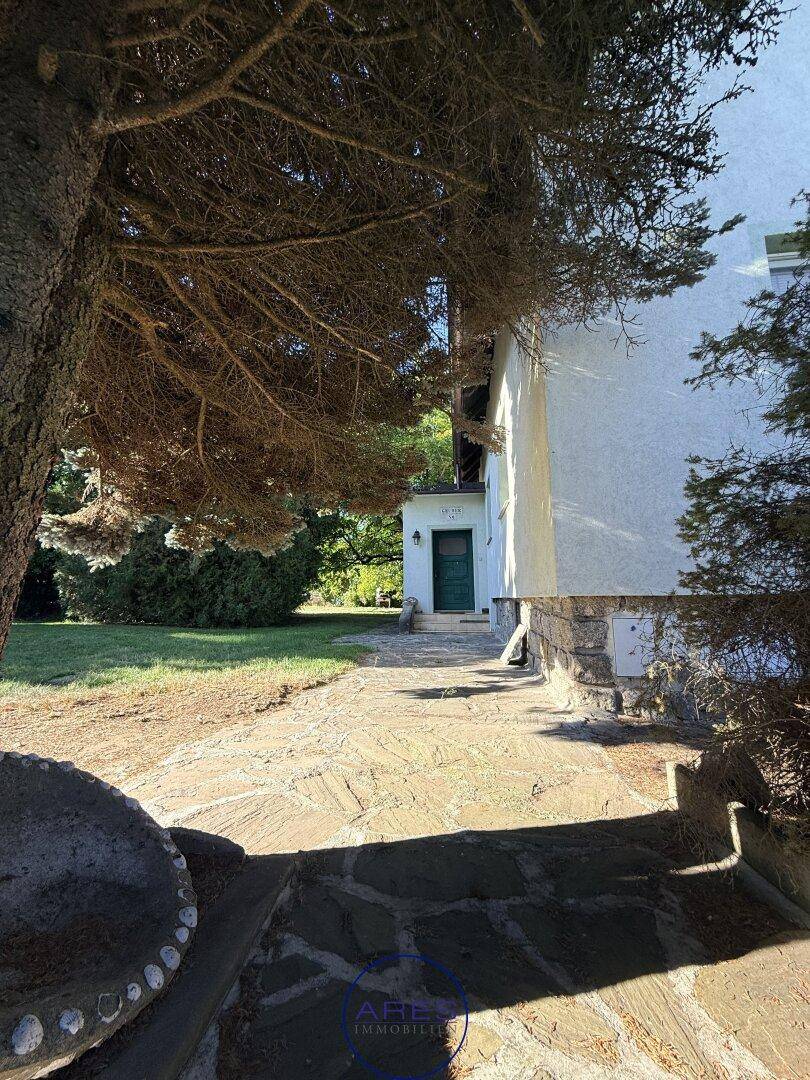 Steinplattenweg führt zum Eingang eines weißen Hauses, umgeben von hohen Bäumen und Grünflächen.