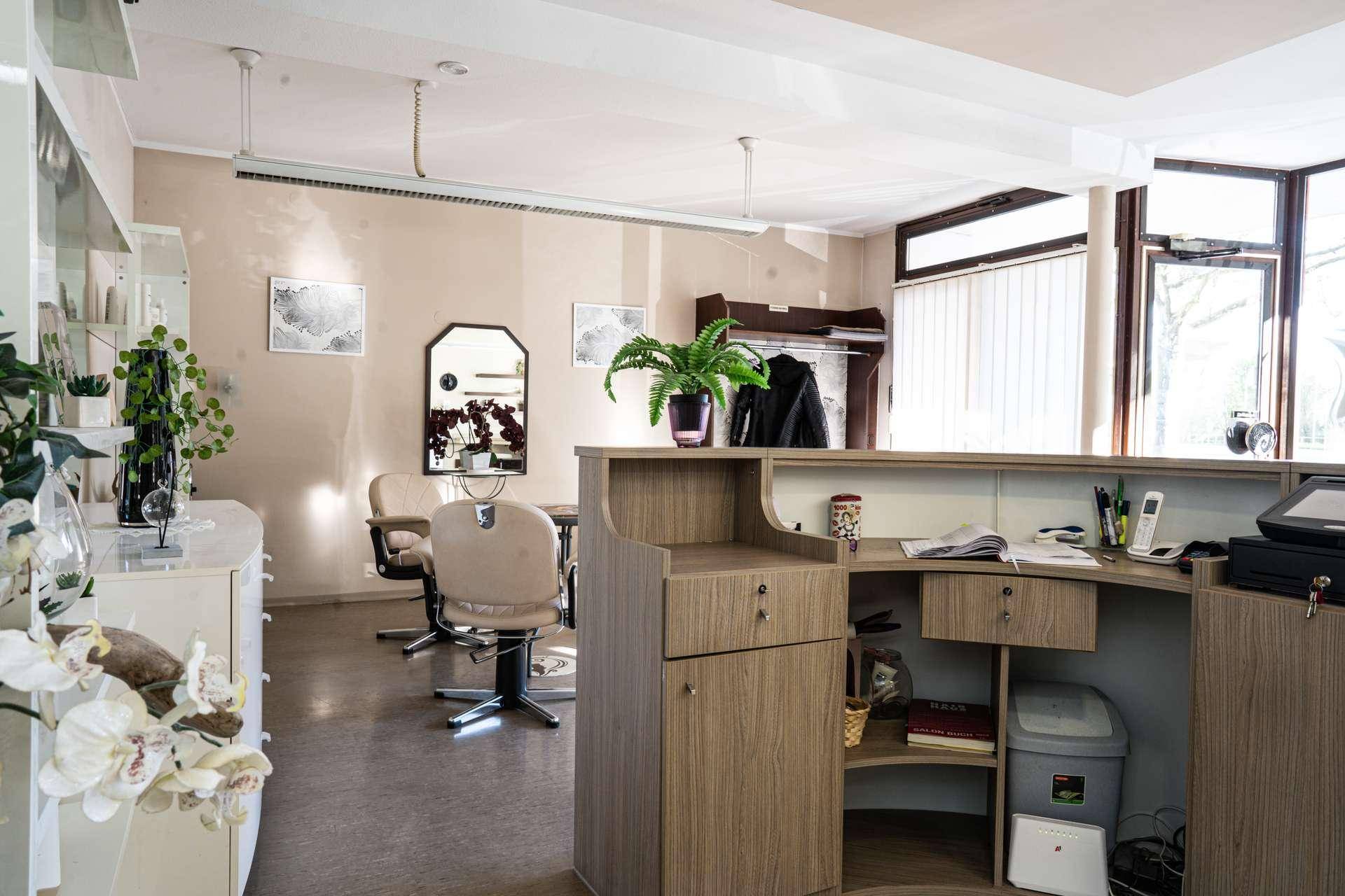 Empfangstheke aus hellem Holz in einem Salon mit Blick auf den Wartebereich und große Fenster.