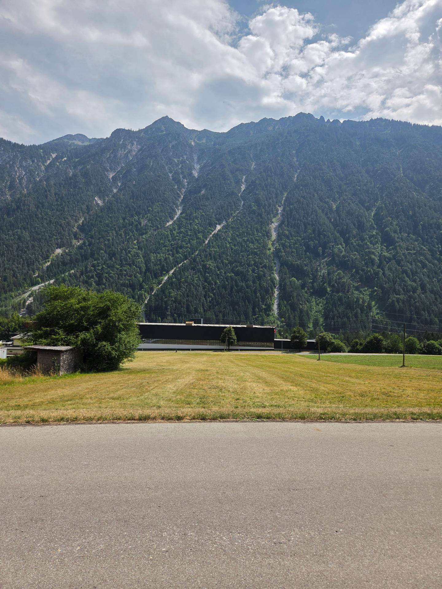 Blick über eine Straße auf ein Feld mit Bergkulisse und einem Gebäude am Horizont unter leicht bewölktem Himmel.