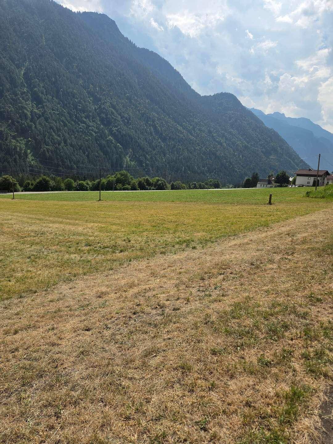 Offenes Feld mit weitem Bergblick und einem Wohnhaus in der Ferne unter leicht bewölktem Himmel.