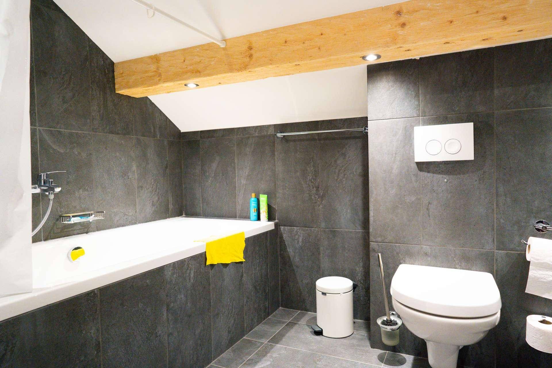Modernes Badezimmer im Dachgeschoss mit Badewanne, dunklen Fliesen und Holzbalken.