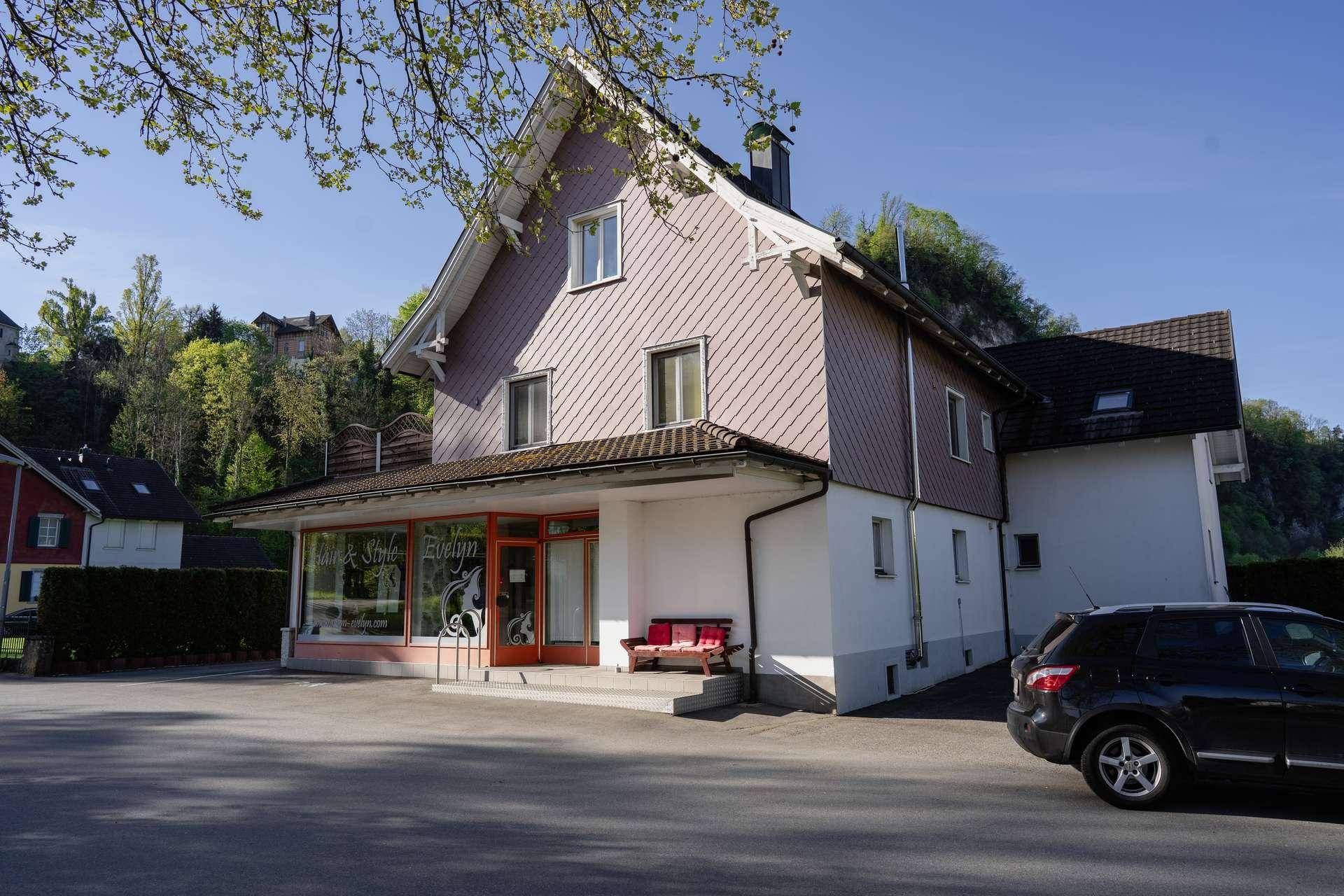 Gebäude mit Giebeldach und Schindelfassade, umgeben von Bäumen und einem Parkplatz unter blauem Himmel.