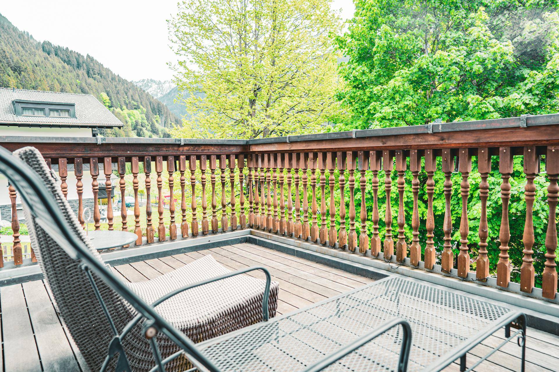 Sonniger Balkon mit Holzgeländer und Liegestuhl, umgeben von grüner Natur.
