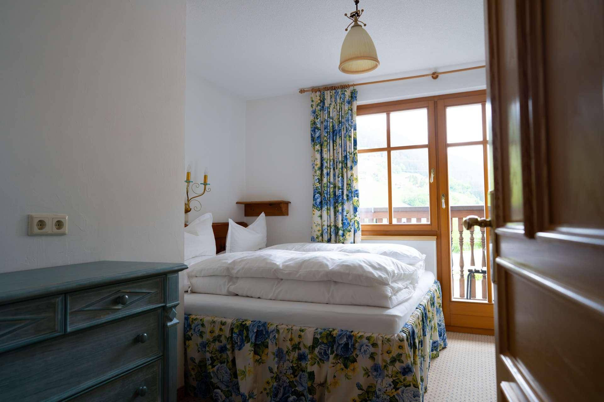 Helles Schlafzimmer mit floralen Vorhängen, Teppichboden und Zugang zum Balkon.