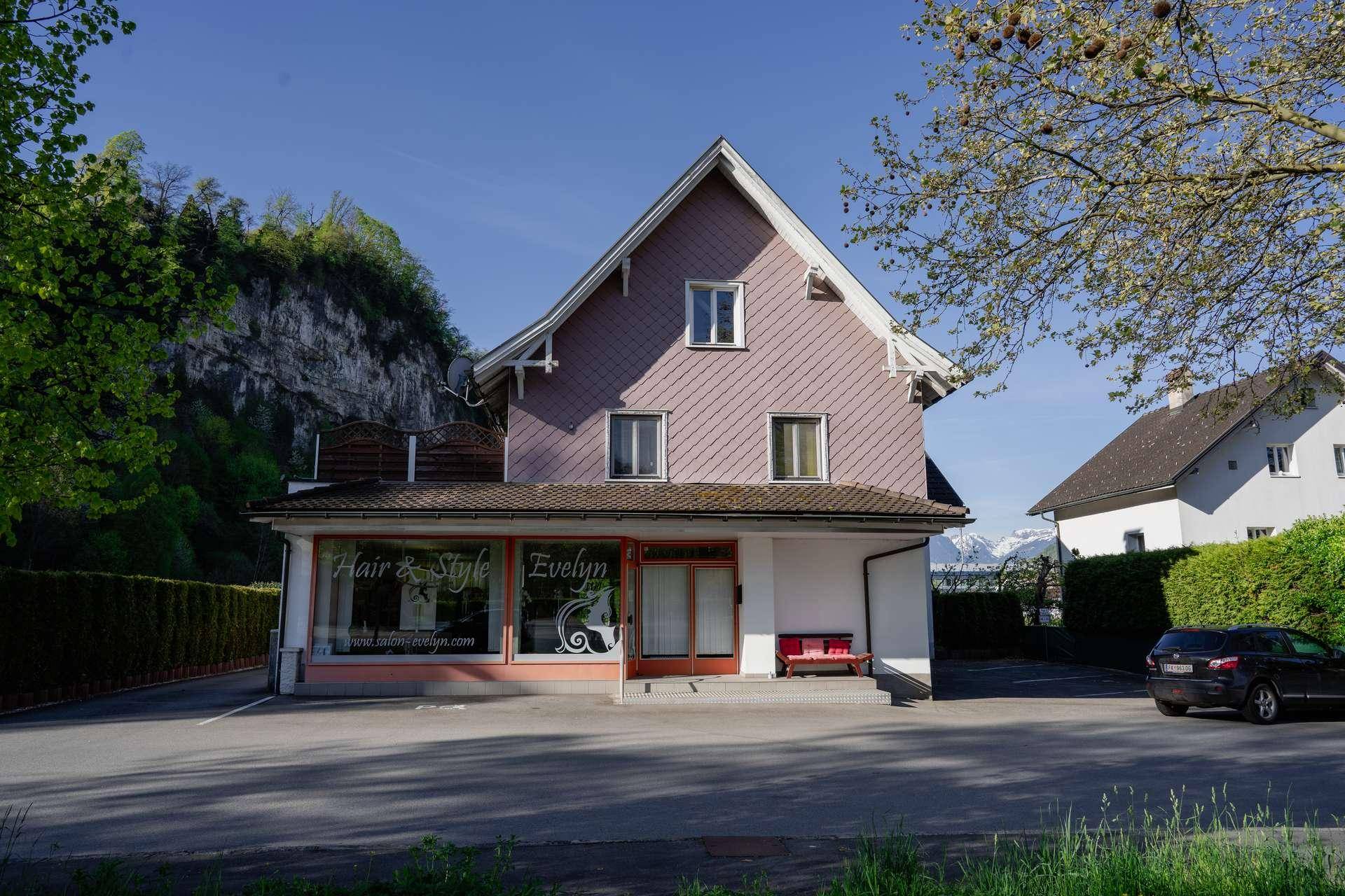 Ansicht des Gebäudes mit Schaufenstern und einem Parkplatz, im Hintergrund sind Berge und grüne Hügel sichtbar.