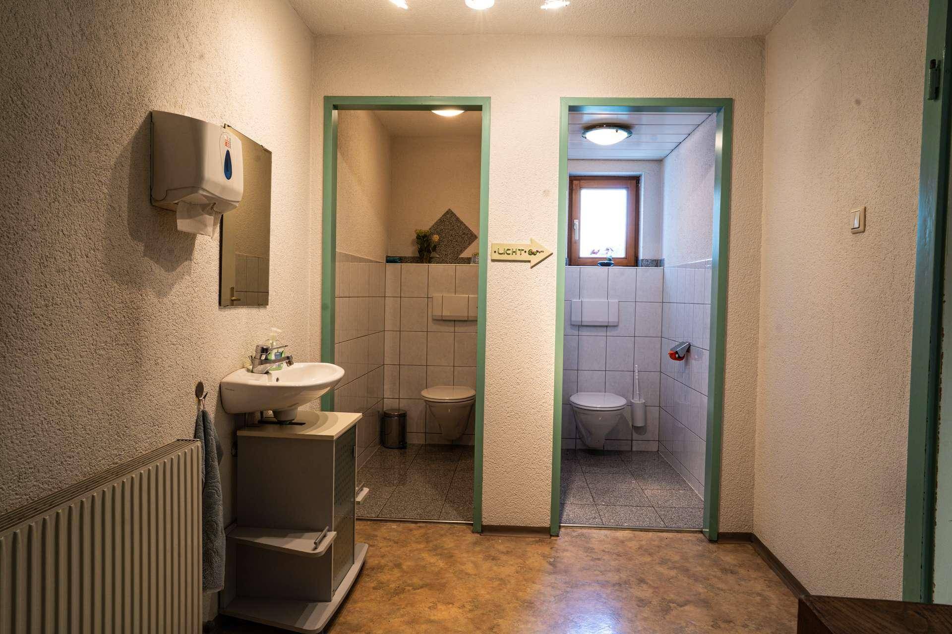 Waschraum mit Waschbecken und zwei separaten Toilettenkabinen, hell gefliest und mit Fenster.