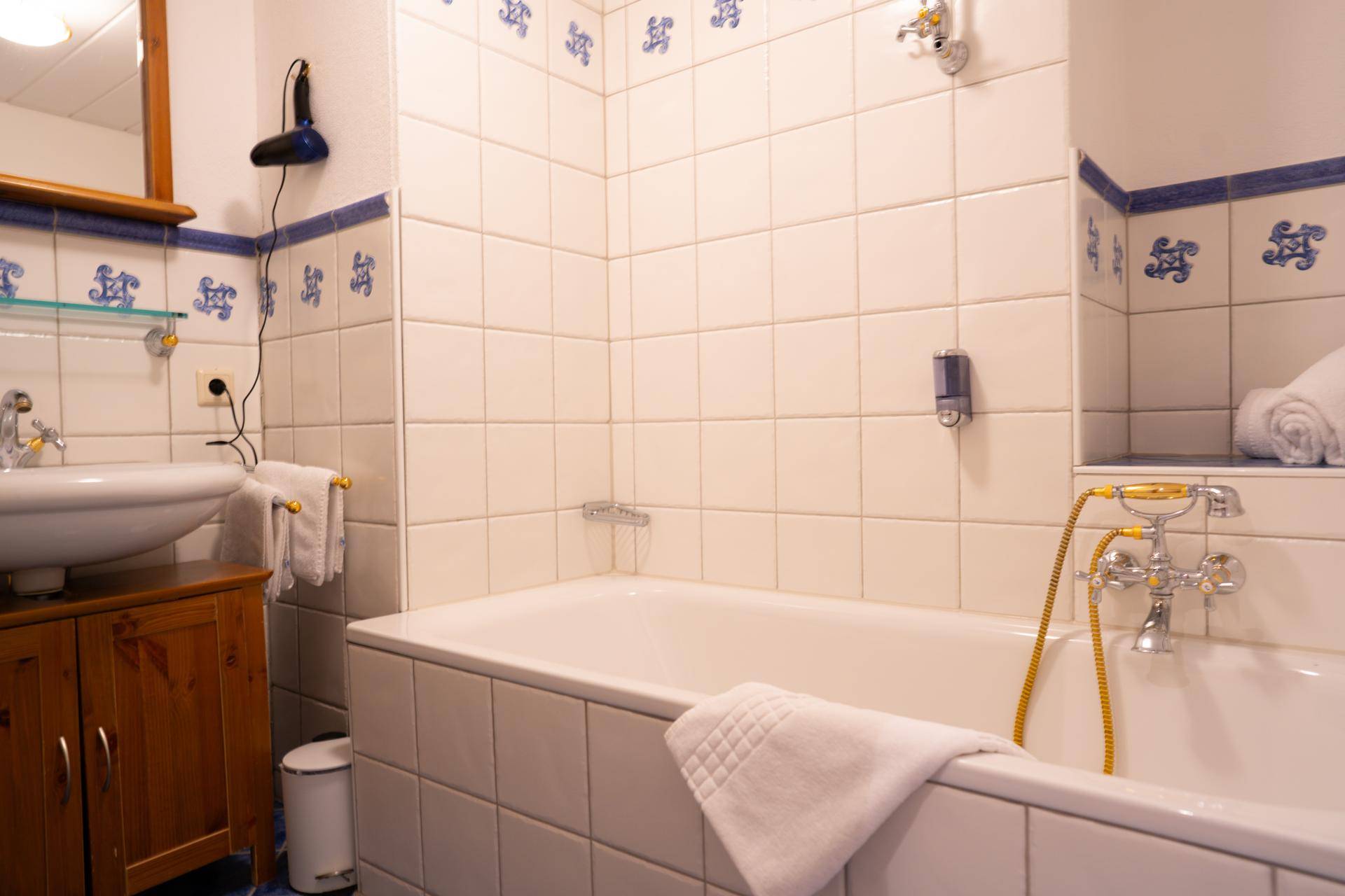 Badezimmer mit Badewanne, Fliesen in Weiß und Blau, sowie einem Waschbecken mit Holzunterschrank.