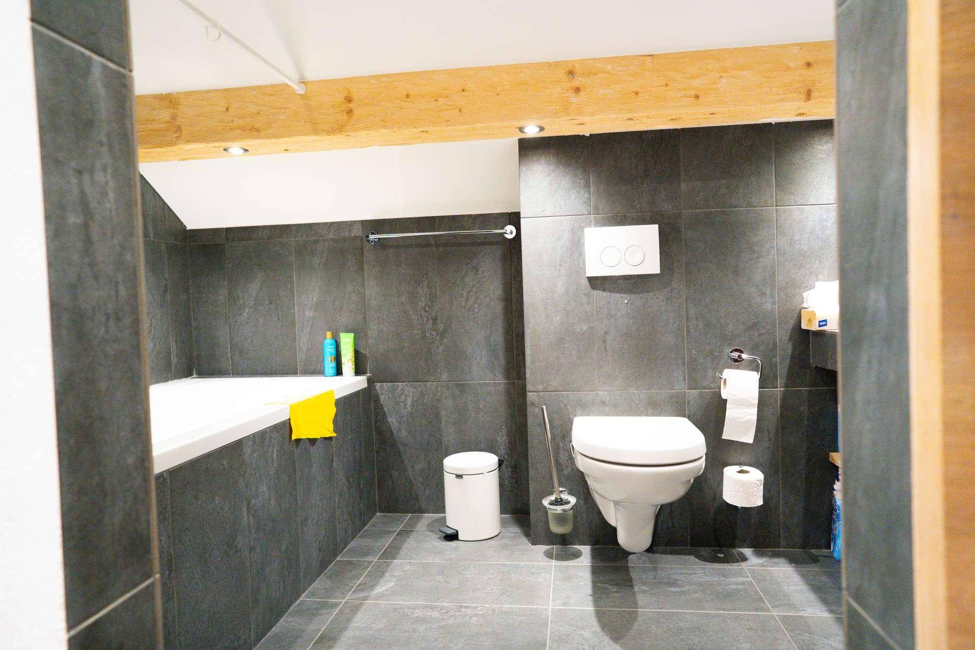 Modernes Badezimmer mit dunklen Fliesen, Badewanne und wandhängendem WC unter sichtbaren Holzbalken.