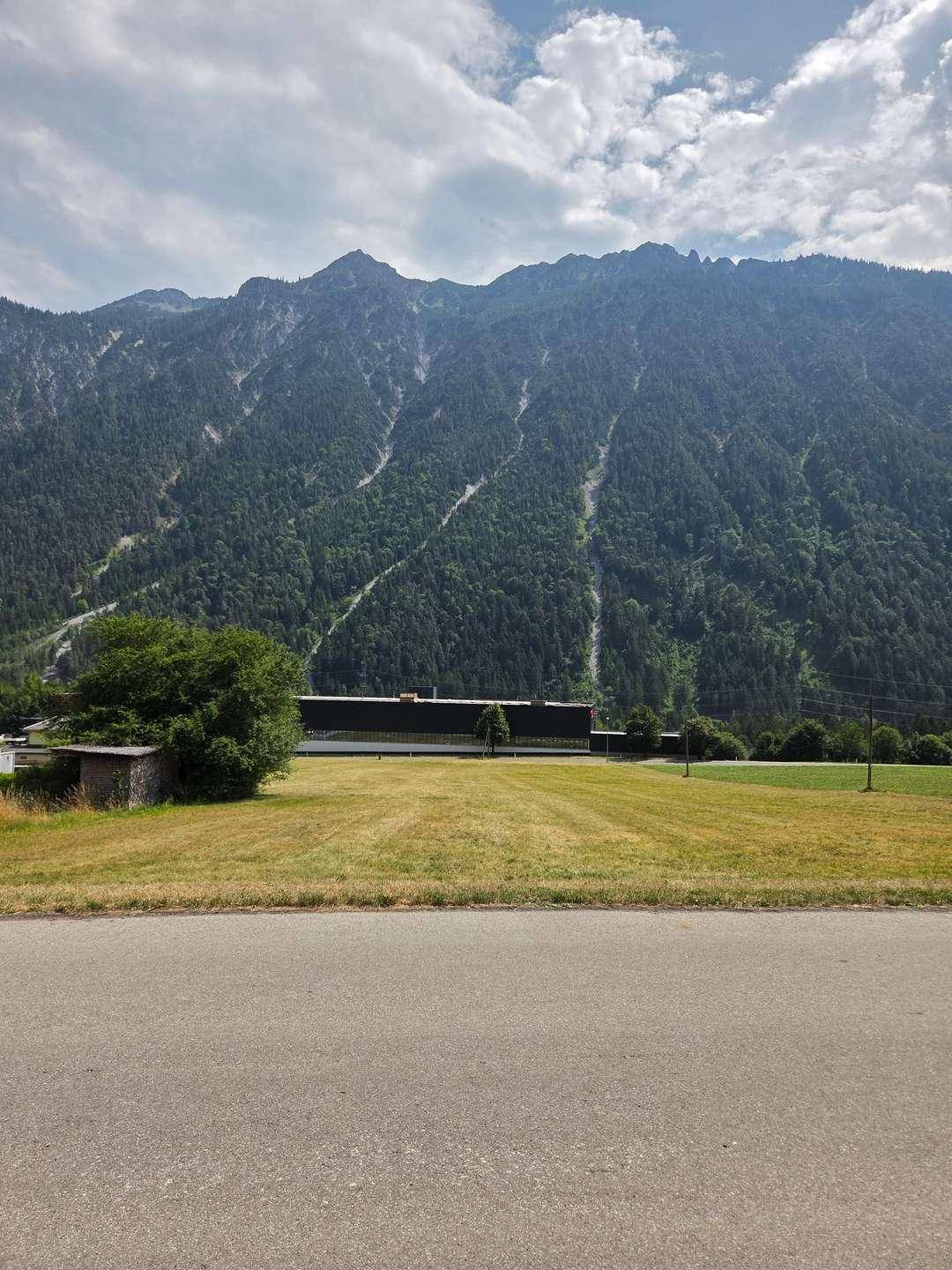 Blick über ein Feld auf die Berge mit einer Straße im Vordergrund und einem Gebäude in der Mitte.