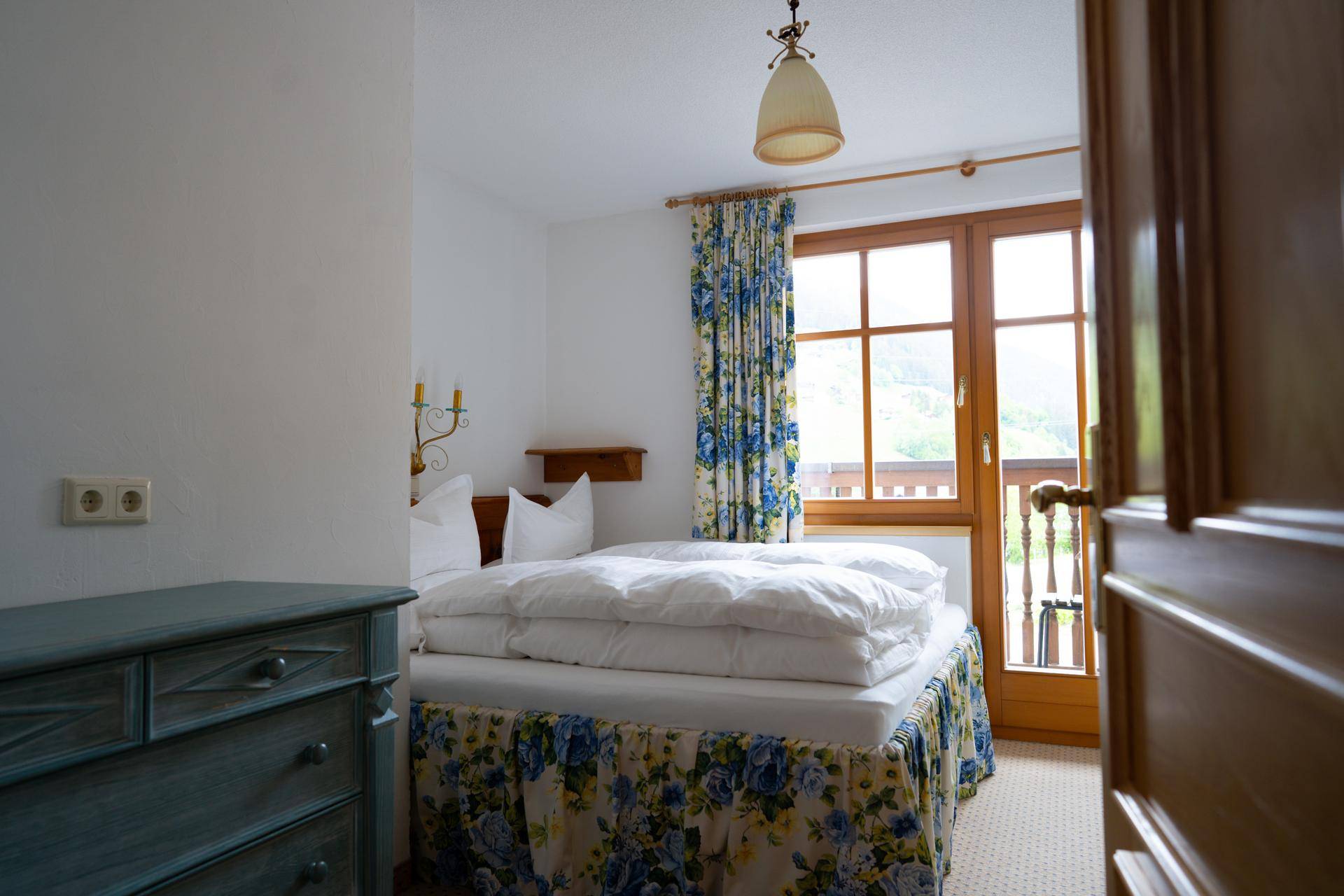 Helles Schlafzimmer mit floralen Vorhängen und Bettwäsche, sowie Zugang zum Balkon.