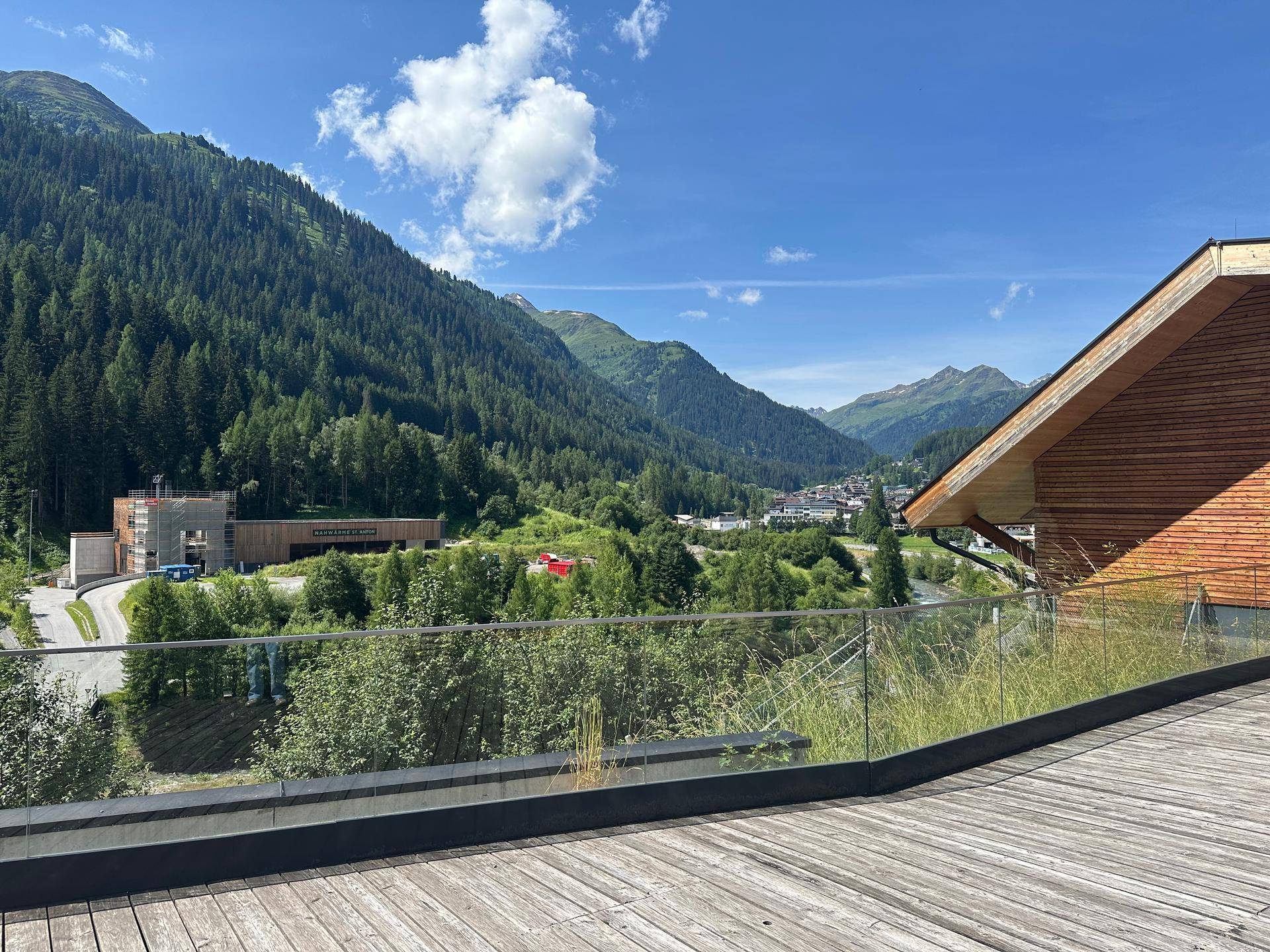 Weitläufige Holzterrasse mit Glasgeländer und Panoramablick auf die umliegende Bergwelt und das Tal.