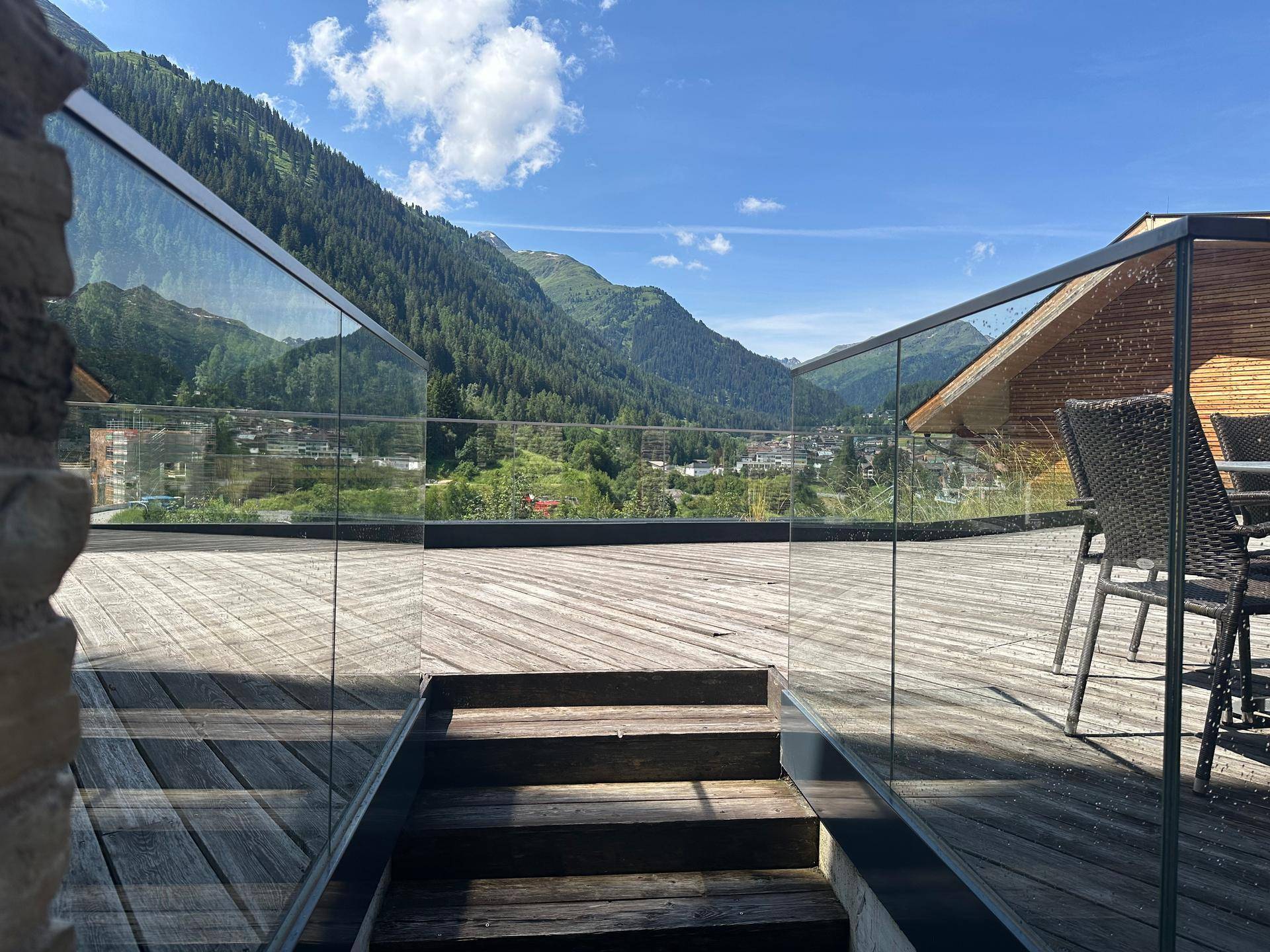 Geräumige Holzterrasse mit Glasgeländer und Treppe, umgeben von einer grünen Berglandschaft.