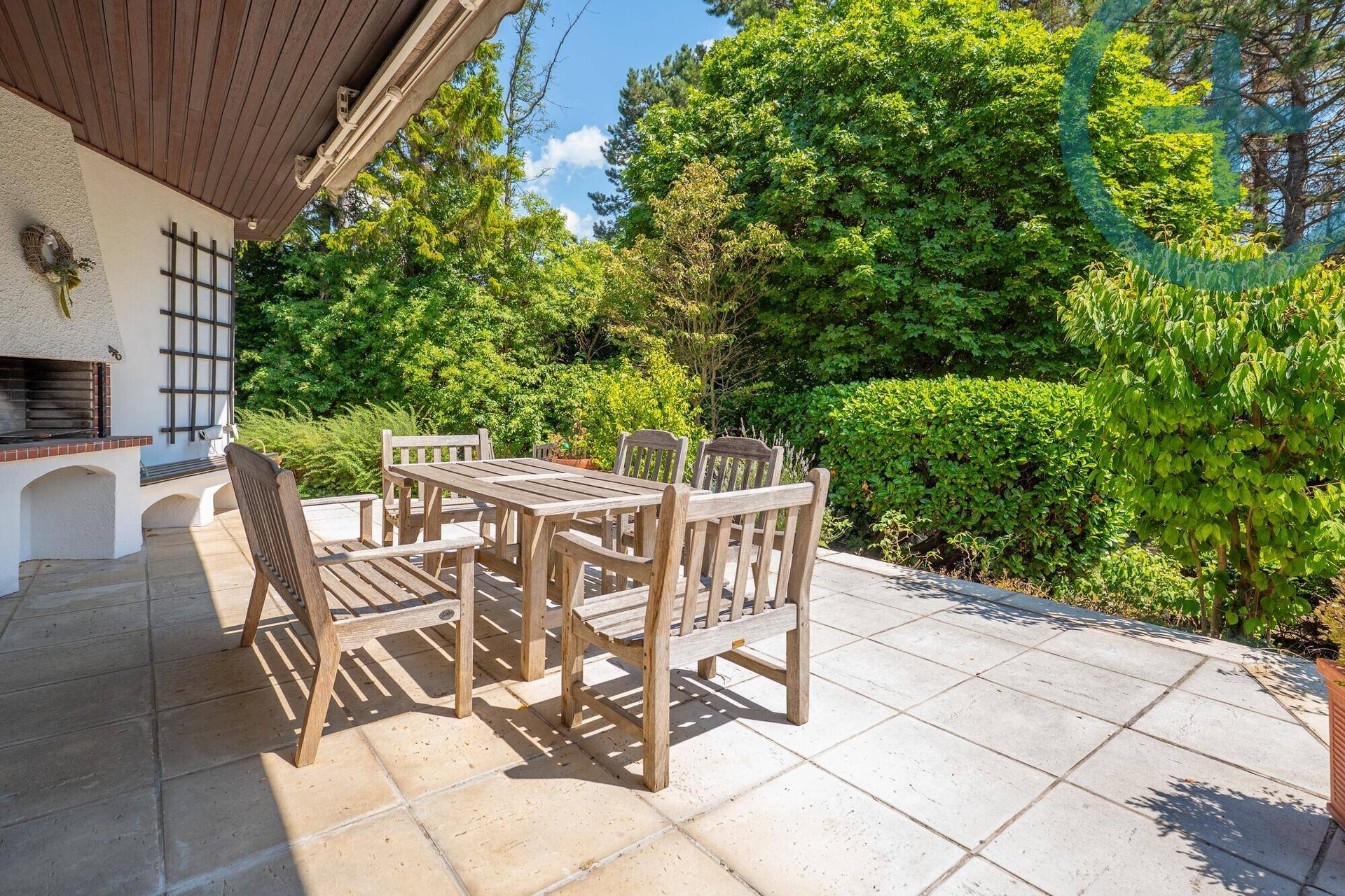 Gemütliche Terrasse mit Holzesstisch, Stühlen und einem integrierten Grillbereich, umgeben von Hecken und Bäumen.