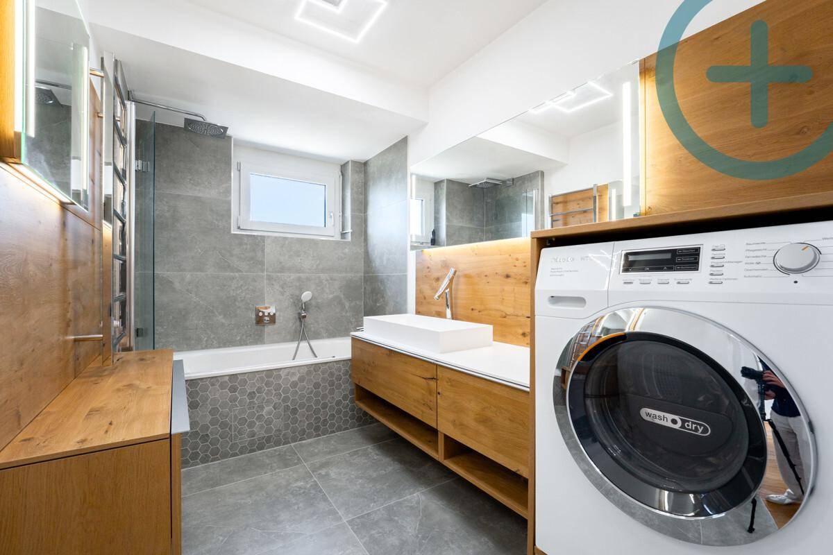 Modernes Badezimmer mit grauen Fliesen, Holzmöbeln, Badewanne, Dusche und Waschmaschine.