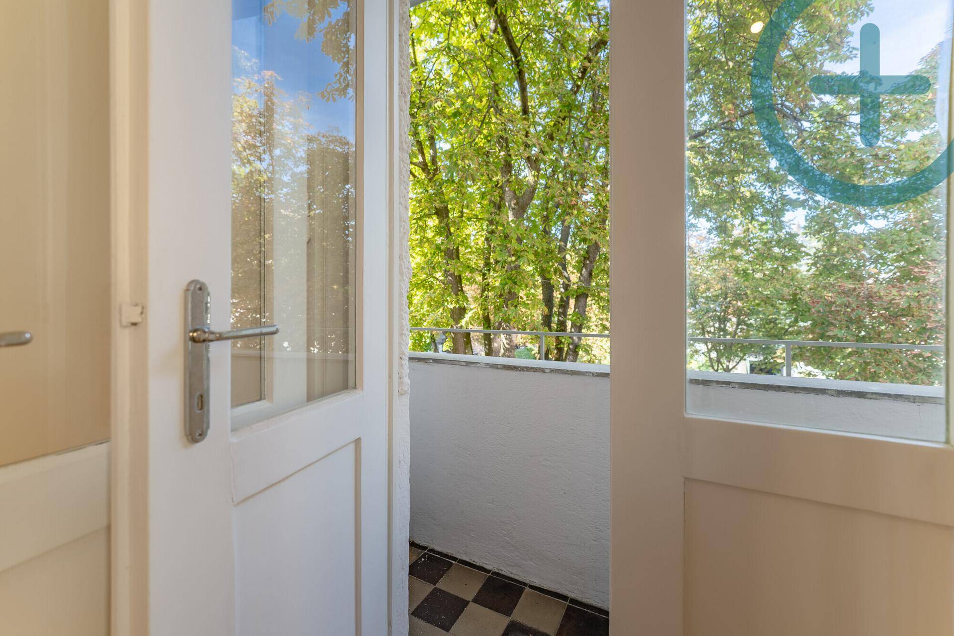 Blick vom Innenraum auf einen kleinen Balkon mit karierten Fliesen und Bäumen.