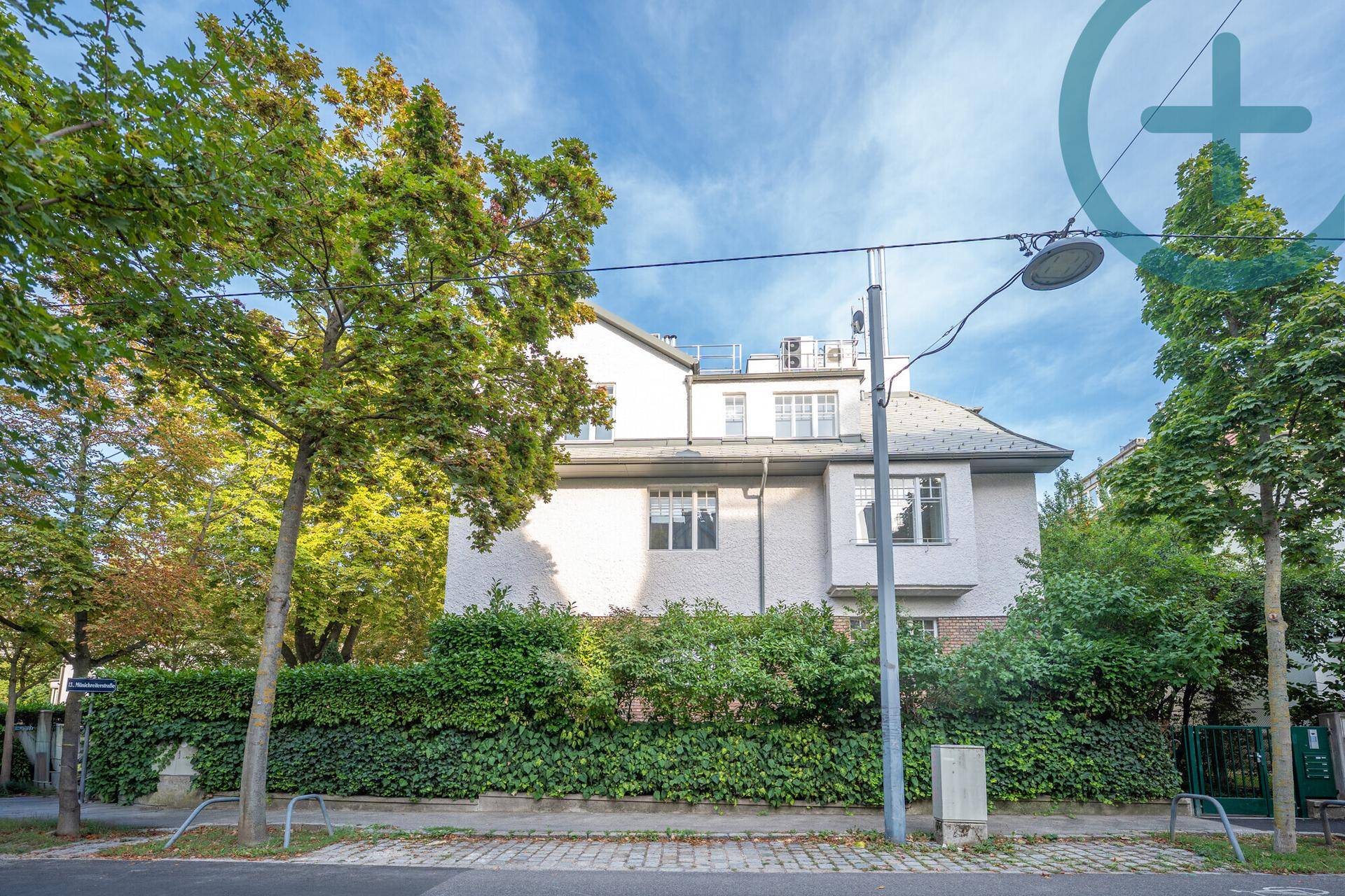 Ansicht eines großen Wohnhauses mit weißer Fassade, umgeben von Bäumen und Hecken.