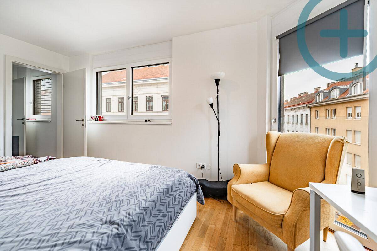 Geräumiges Schlafzimmer mit großem Fenster, Sessel und Blick auf die umliegenden Gebäude.