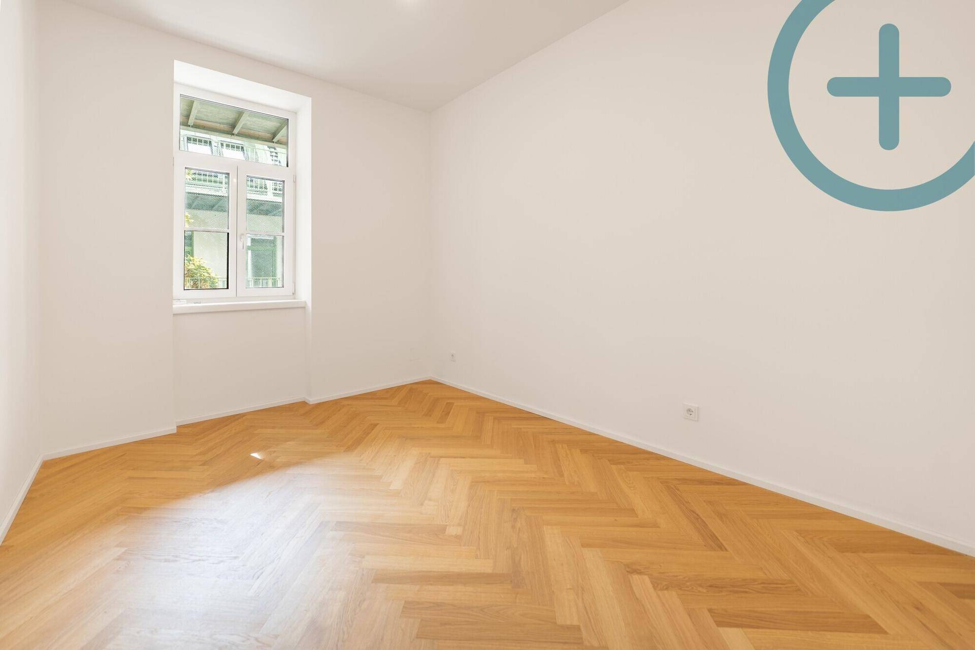 Helles Schlafzimmer mit Fischgrätparkett und einem großen Fenster, das Tageslicht spendet.