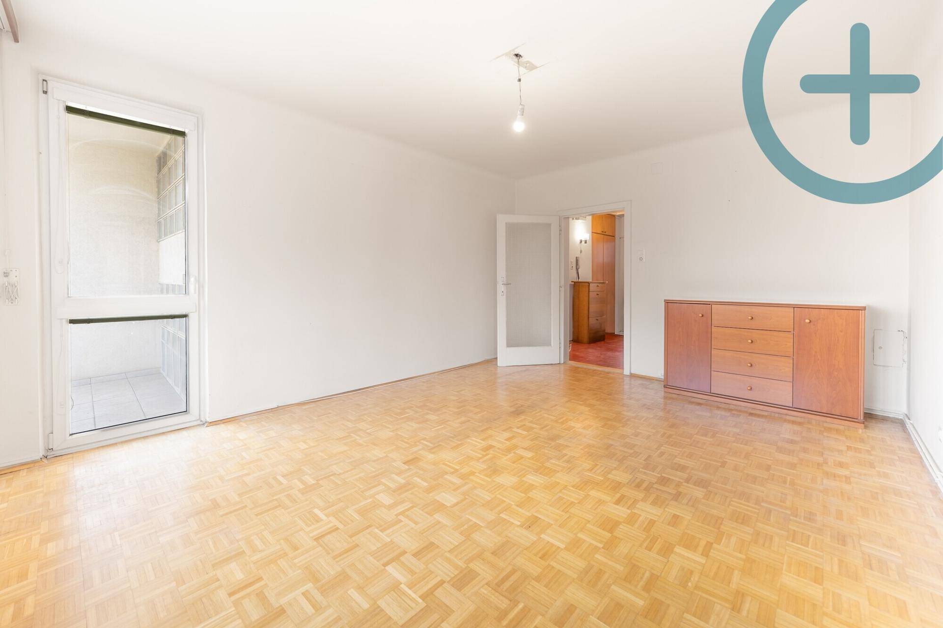 Geräumiges Wohnzimmer mit Fischgrätparkett, hellen Wänden und einem Holzschrank.