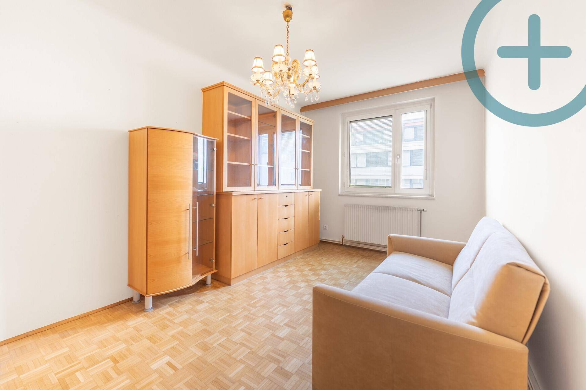 Wohnzimmer mit Parkettboden, Holzvitrine, Sofa und Kronleuchter, hell durch großes Fenster.