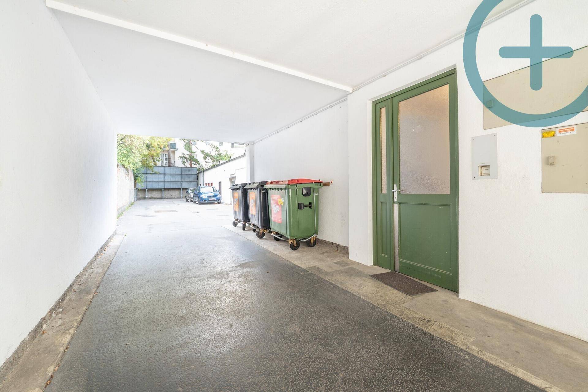 Überdachter Außenbereich mit Mülltonnen und Zufahrt zu einem Parkplatz.