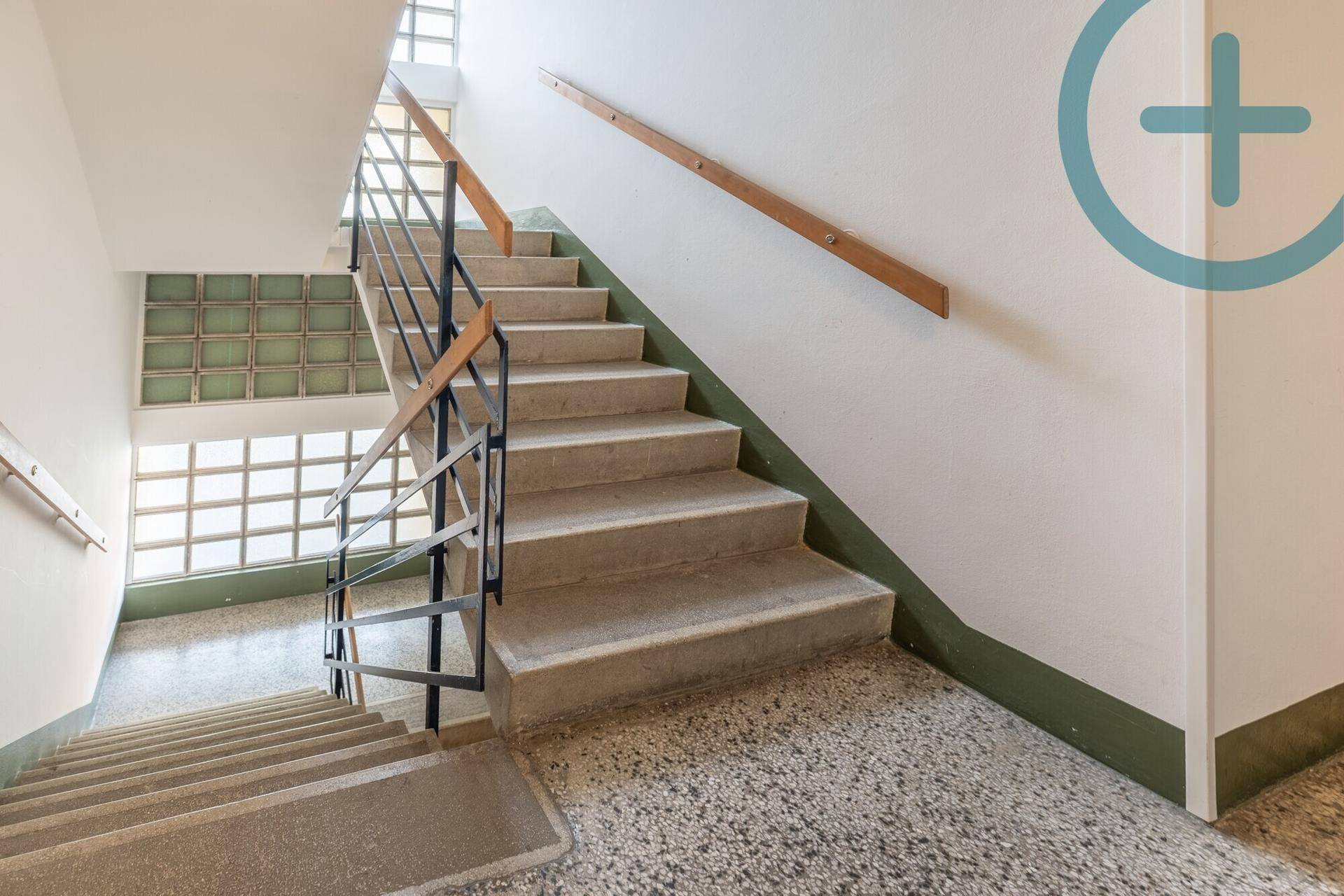 Treppenhaus mit Betonstufen, Metallgeländer und Glasbausteinfenstern im Gemeinschaftsbereich.