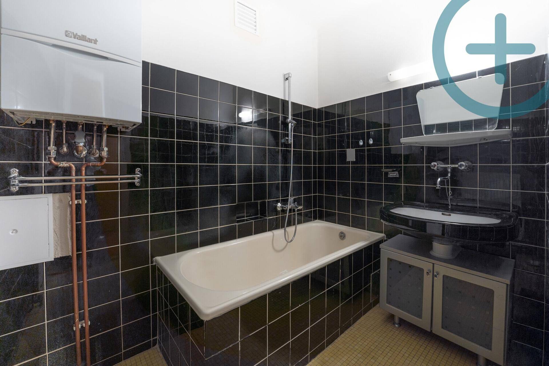 Badezimmer mit schwarzen Fliesen, Badewanne mit Duschfunktion und Warmwasserboiler.