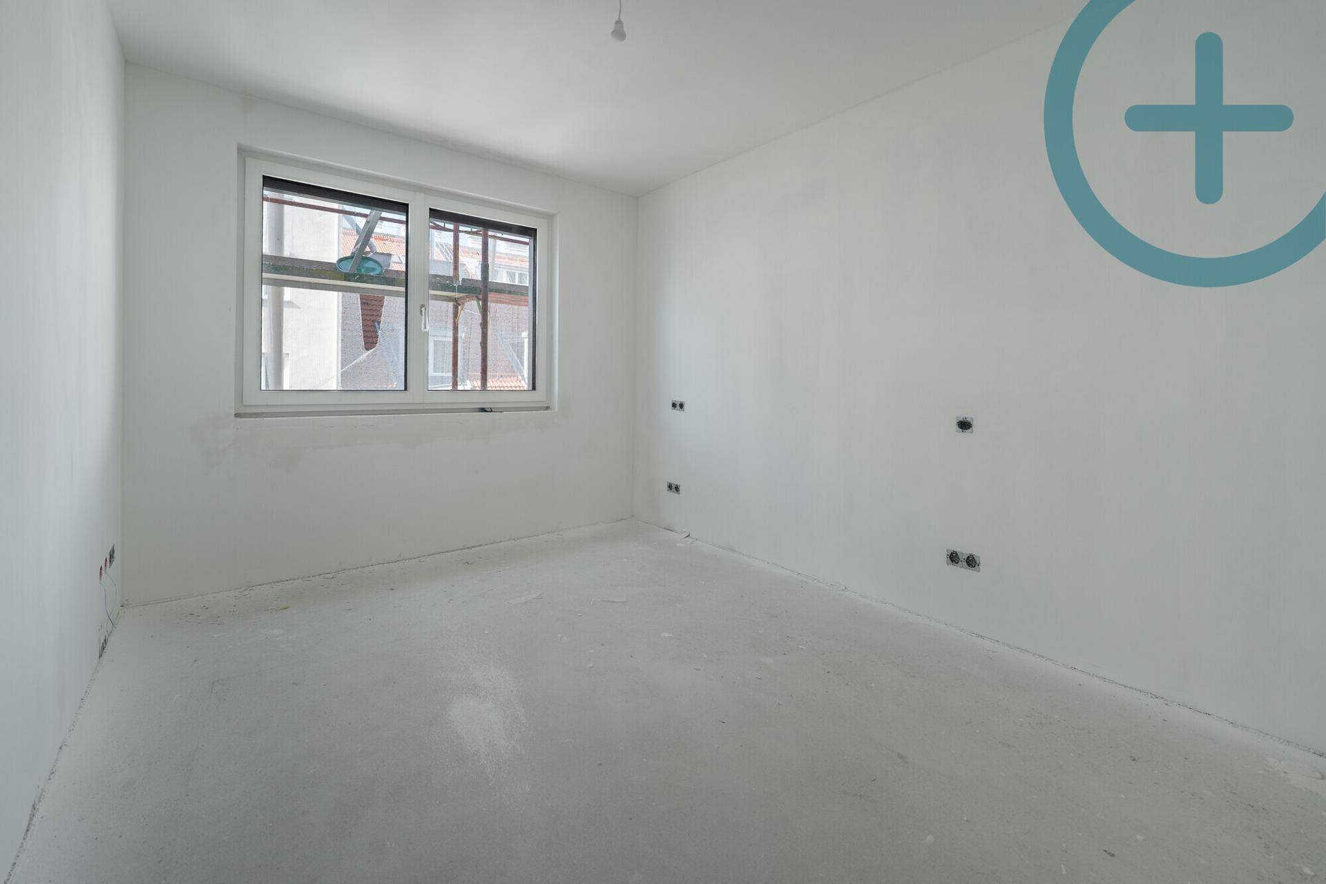 Leerer Raum im Rohbau mit Betonboden, weißen Wänden und einem Fenster für Tageslicht.