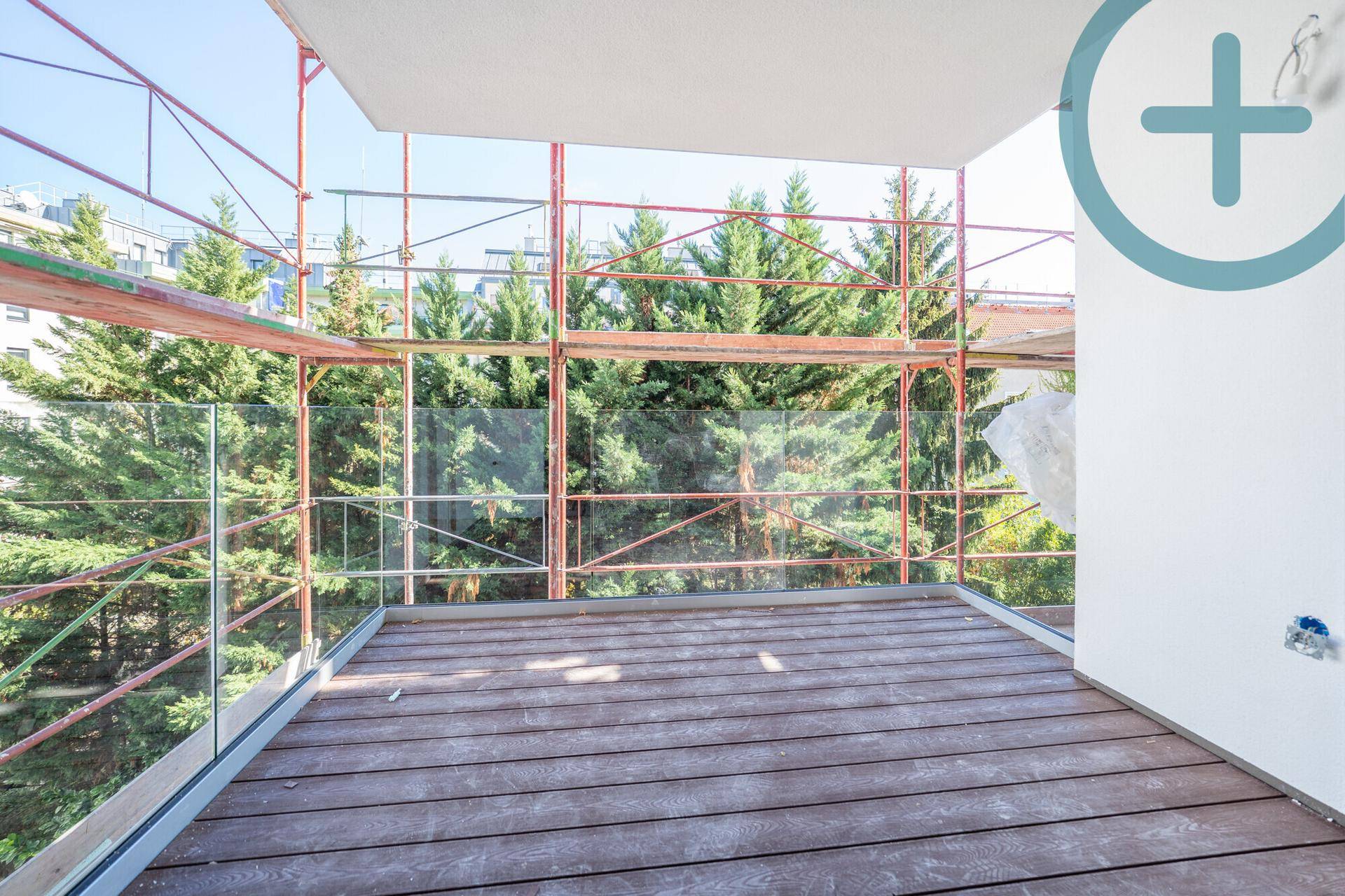 Balkon mit Holzbelag und Glasgeländer, umgeben von Bäumen und Baugerüst im Hintergrund.