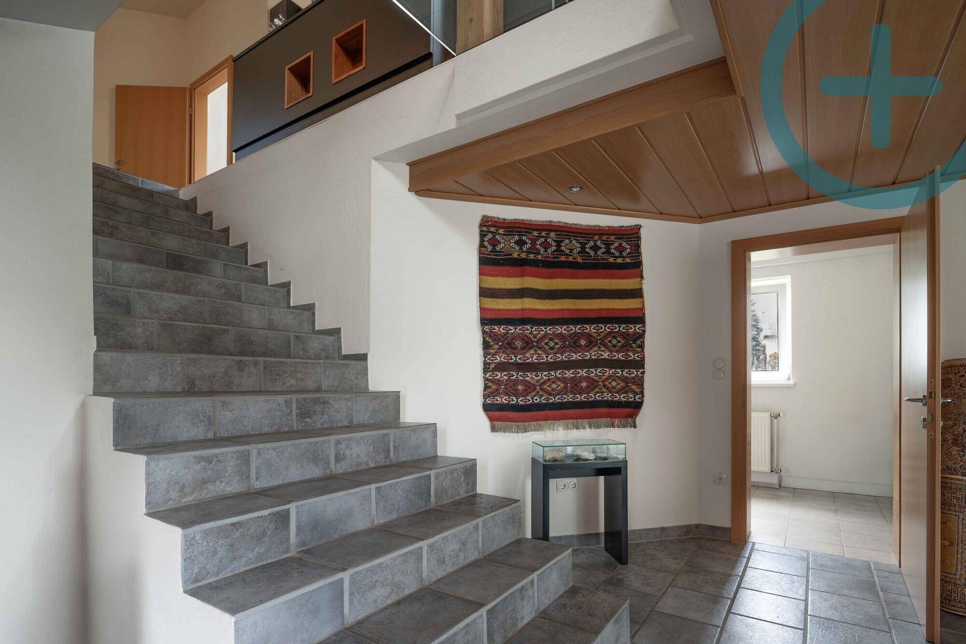 Heller Eingangsbereich mit Steintreppe, Holztäfelung an der Decke und dekorativem Wandteppich.