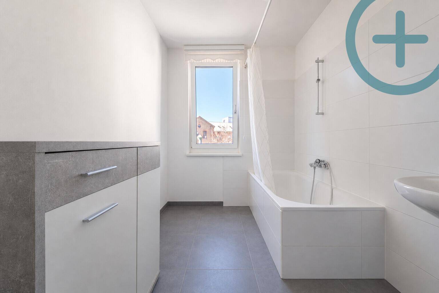 Modernes Badezimmer mit Badewanne, Waschbecken, Fenster und grauen Bodenfliesen.