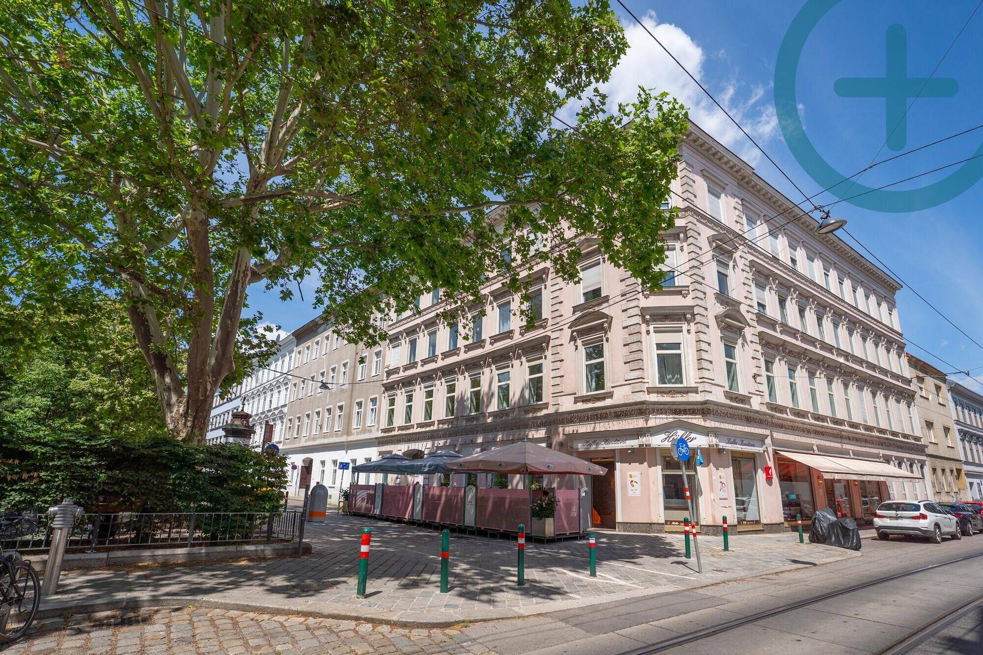 Eckgebäude mit klassischer Architektur, Bäumen und Straßenbahnschienen im Vordergrund.