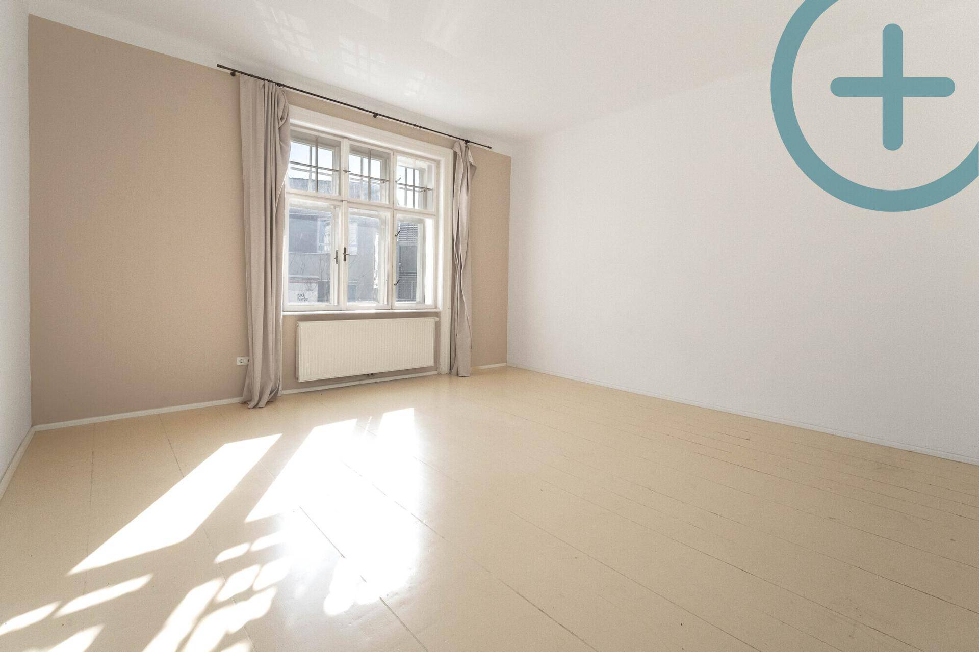 Heller, leerer Raum mit Holzboden und einem großen Fenster, das viel Licht hereinlässt.
