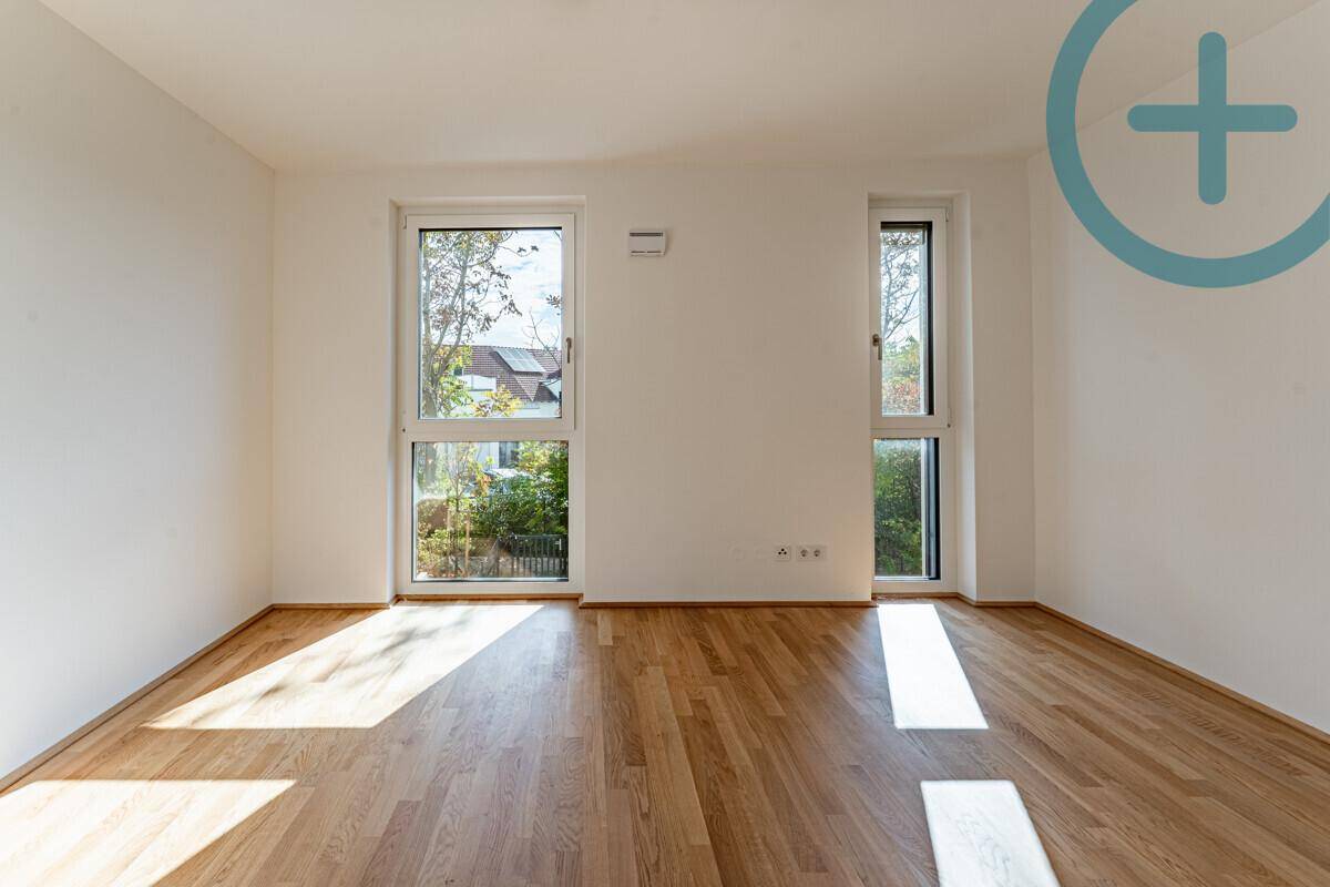 Lichtdurchflutetes Zimmer mit Holzboden und großen Fenstern, die einen Blick ins Grüne bieten.