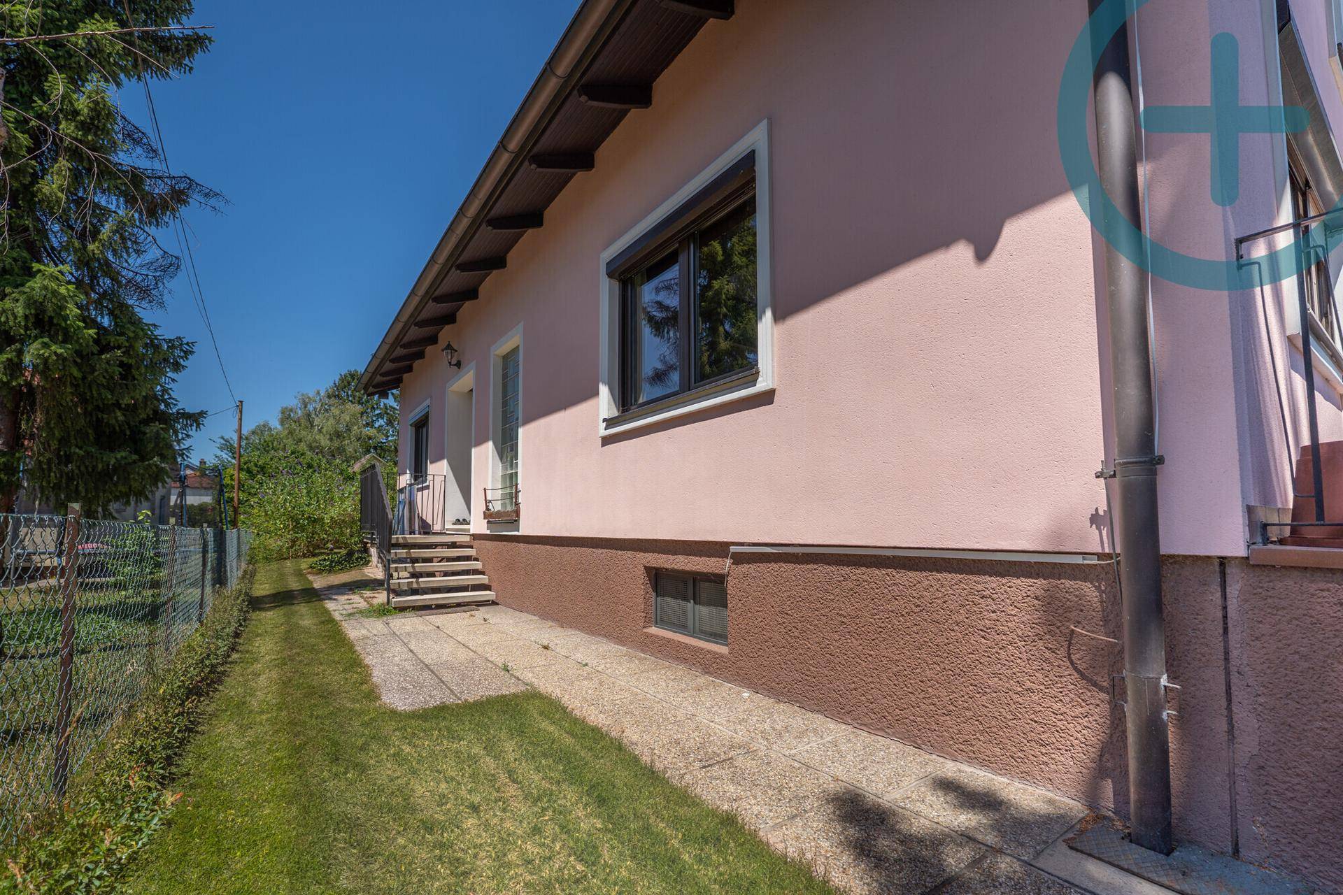 Seitenansicht des Hauses mit hellrosa Fassade, Fenstern und einem schmalen Zugangsweg.