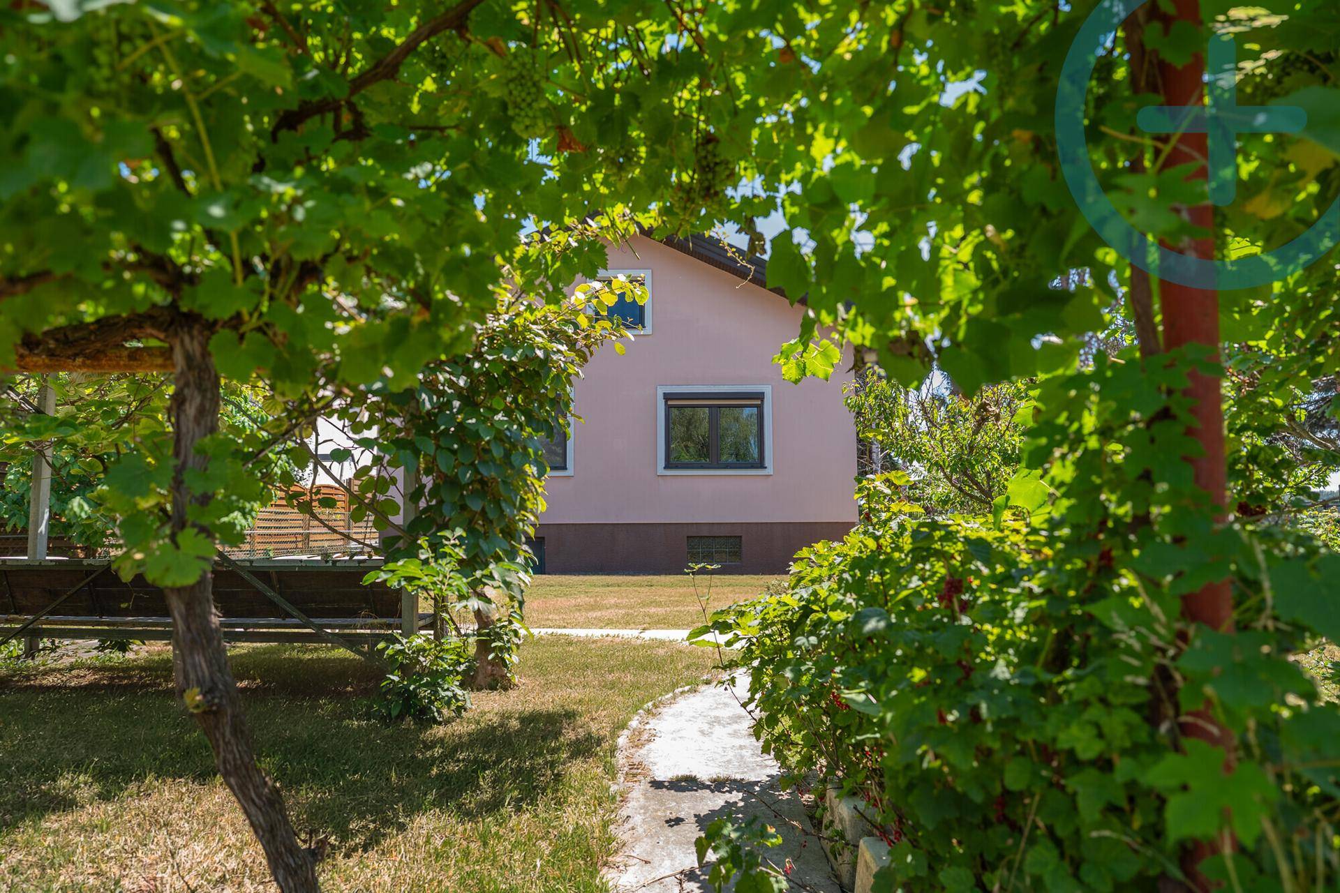 Ein schmaler Gartenweg führt durch üppiges Grün zum hellrosa gestrichenen Haus mit dunklen Fenstern.