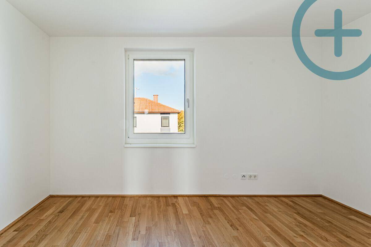 Helles Zimmer mit Parkettboden und einem Fenster, das einen Blick auf die umliegenden Häuser bietet.
