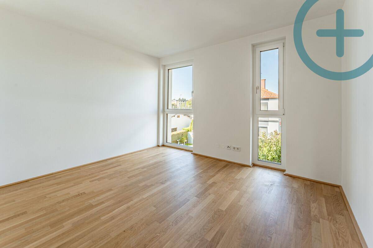 Helles Zimmer mit Parkettboden und zwei hohen Fenstern, die viel Tageslicht hereinlassen.