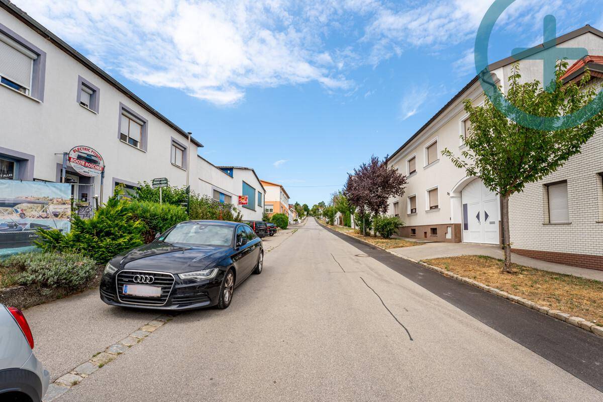 Ruhige Wohnstraße mit geparkten Autos, weißen Häusern und Bäumen am Straßenrand.