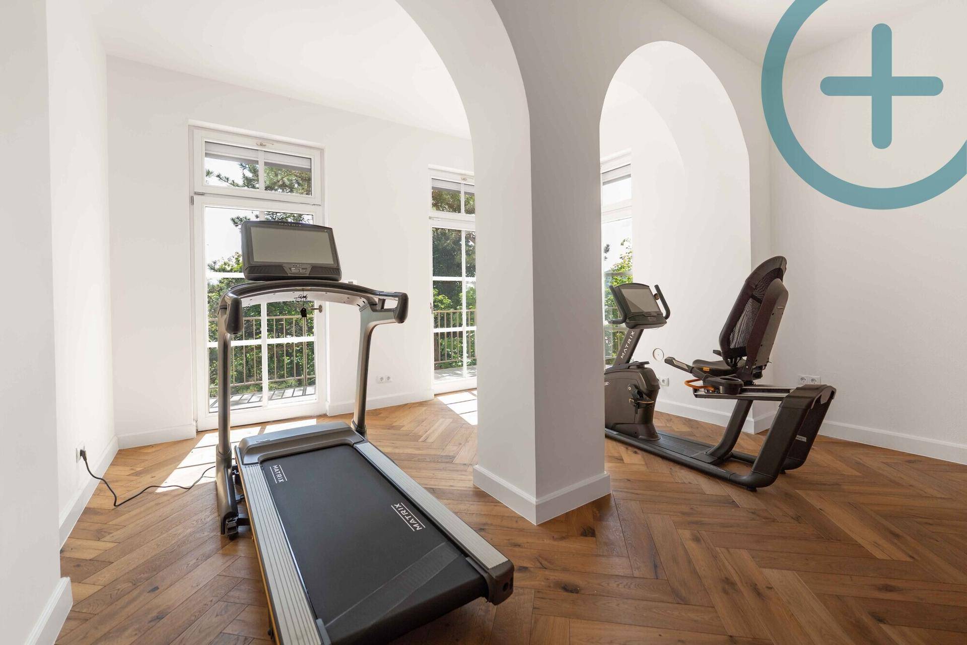 Fitnessraum mit Trainingsgeräten, Fischgrätparkett und großen Fenstern mit Grünblick.
