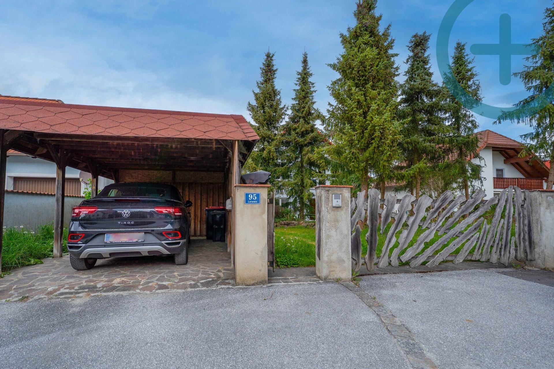 Überdachter Carport mit geparktem Auto, umgeben von hohen Bäumen und einem Holzzaun.