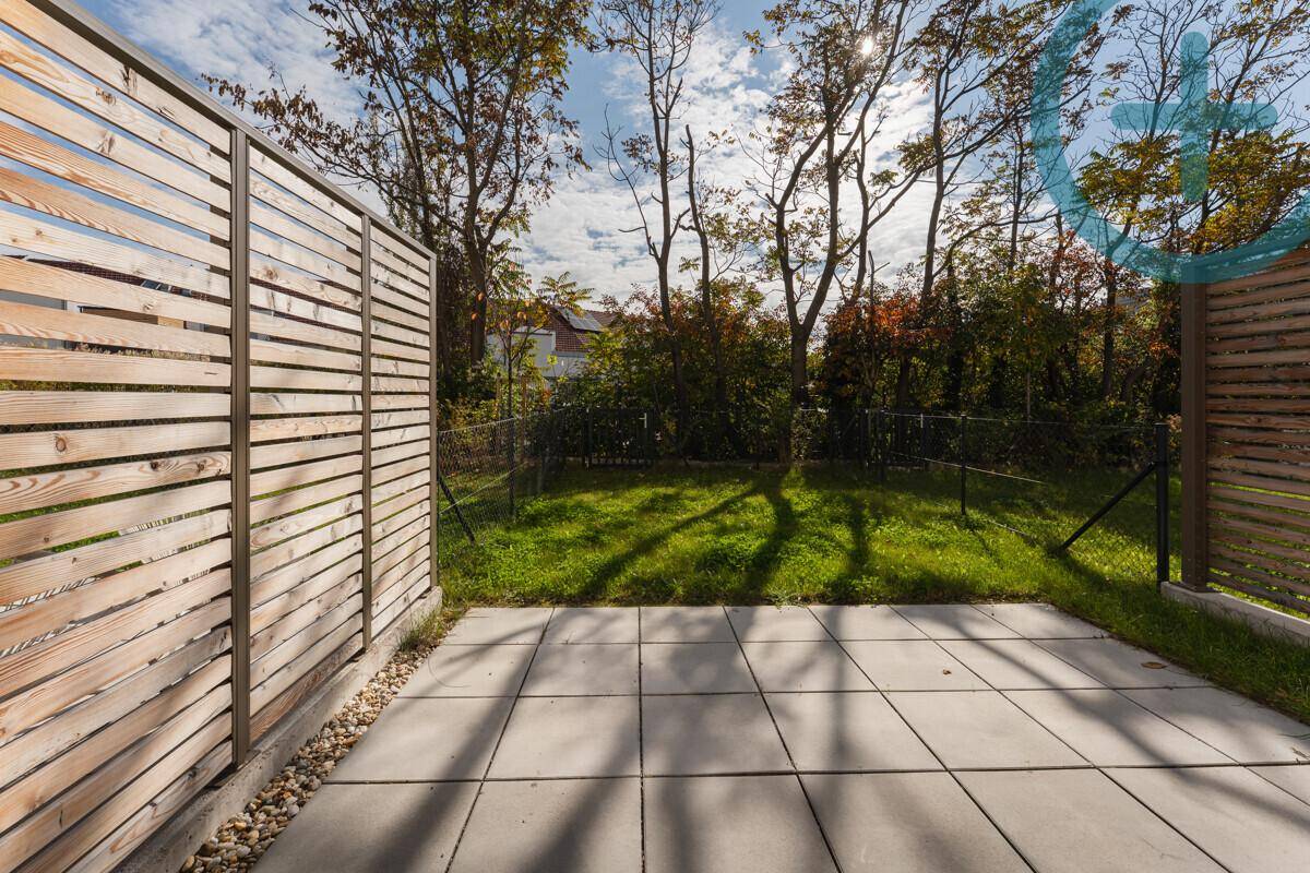 Sonniger Terrassenbereich mit Steinplatten und angrenzendem Rasen, umgeben von einem Holzzaun.