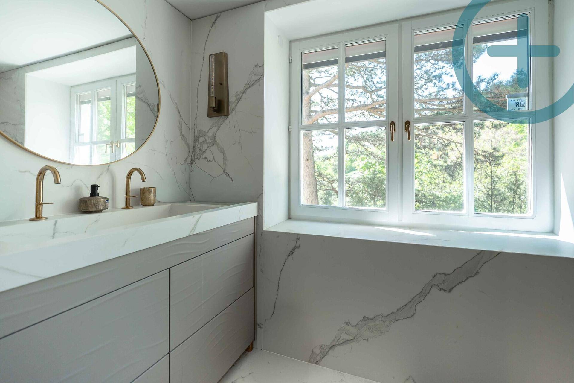 Elegantes Badezimmer mit Marmorwaschtisch, rundem Spiegel und Fenster mit Grünblick.