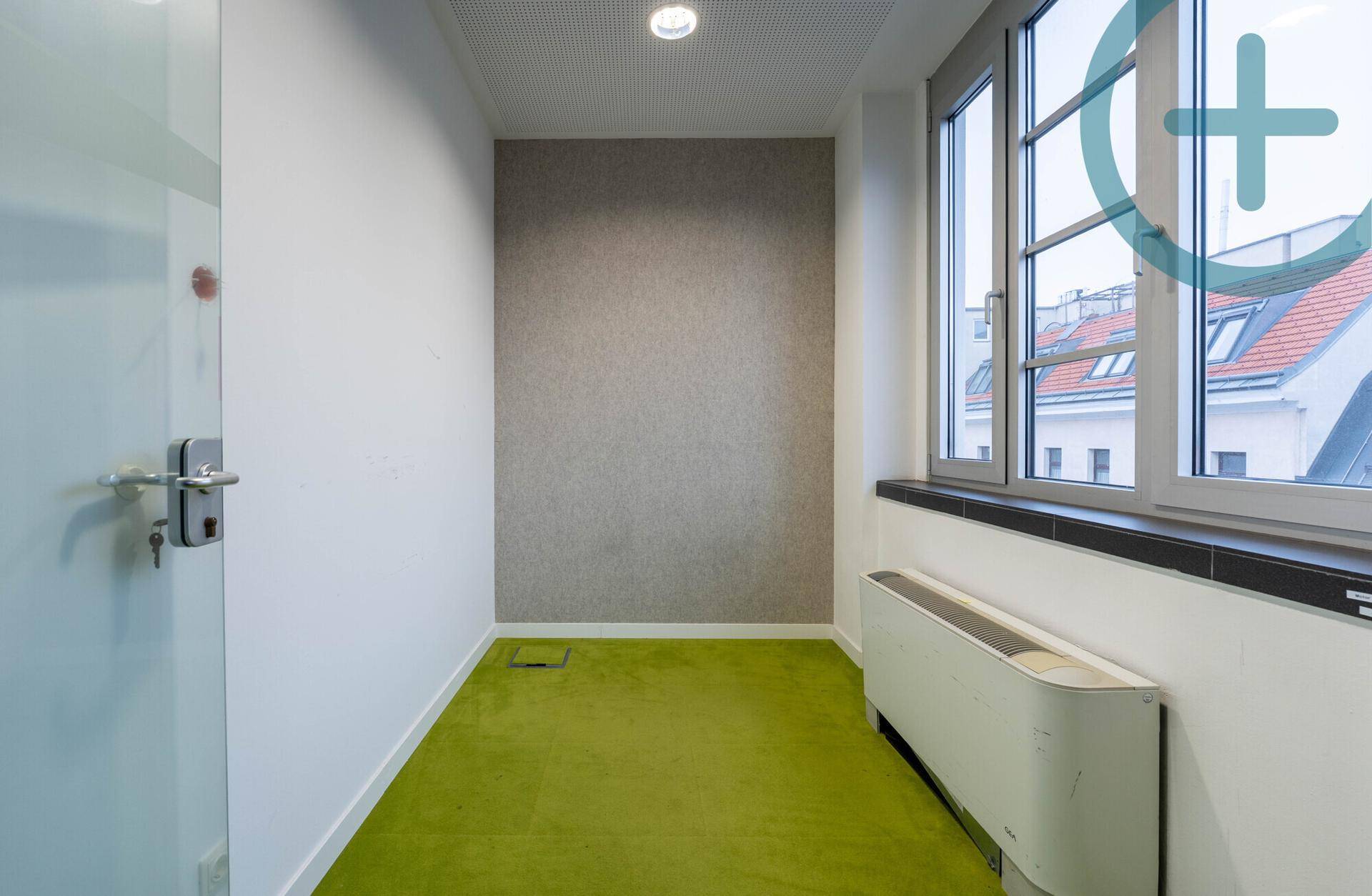 Kleiner, heller Büroraum mit grünem Teppichboden, einer Akzentwand und einem großen Fenster.