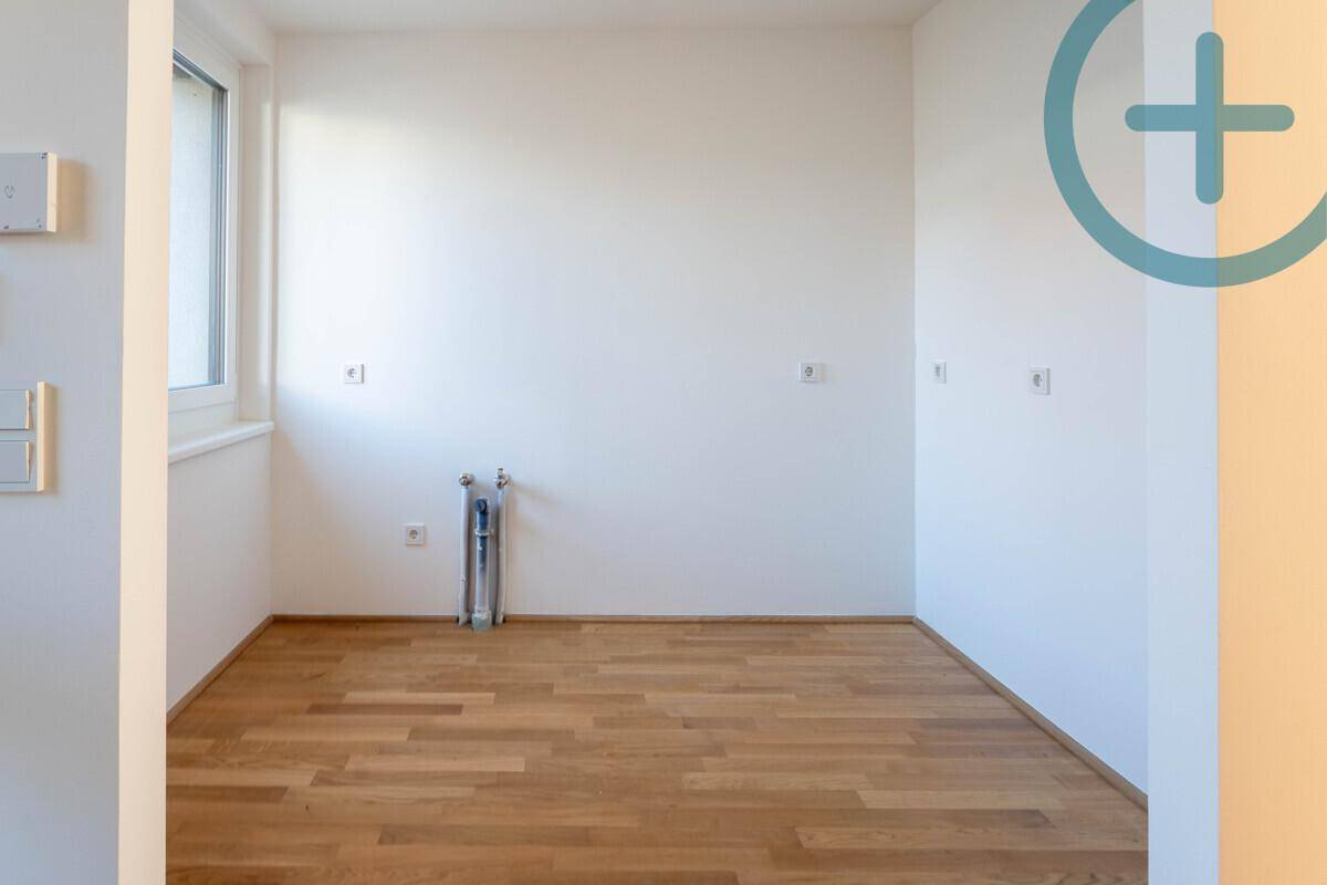 Leerer Küchenraum mit Parkettboden, Fenster und vorbereiteten Anschlüssen für eine Einbauküche.