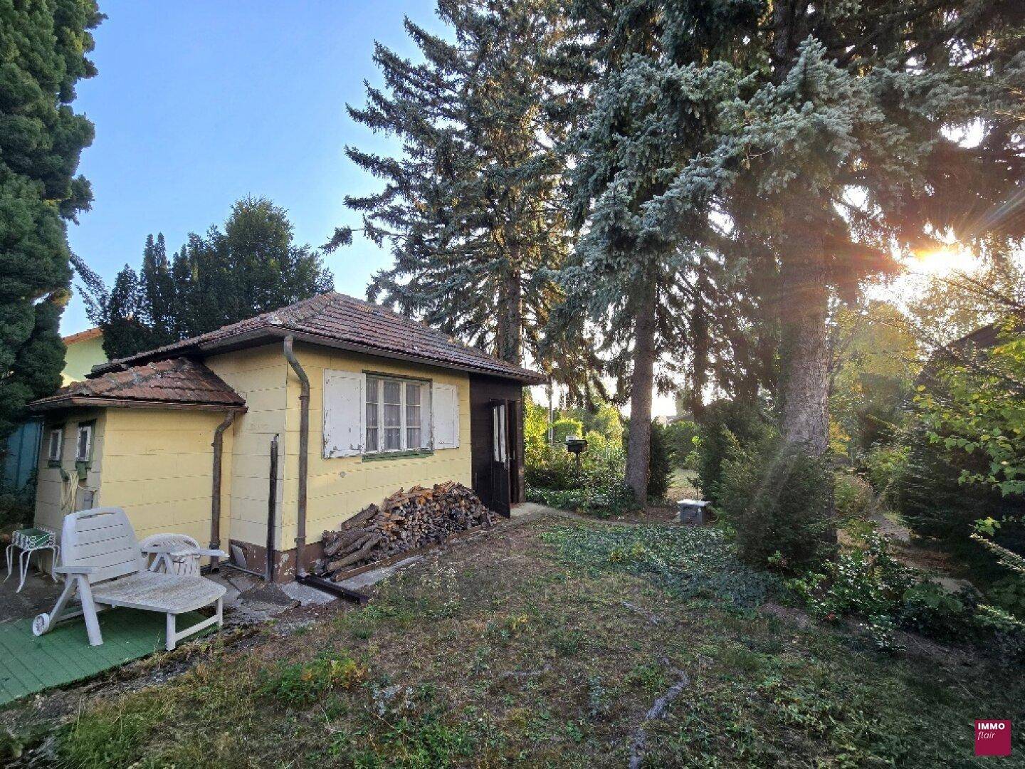 Kleines gelbes Gartenhaus mit Ziegeldach, umgeben von Bäumen und Holzstapeln, bei Sonnenuntergang.